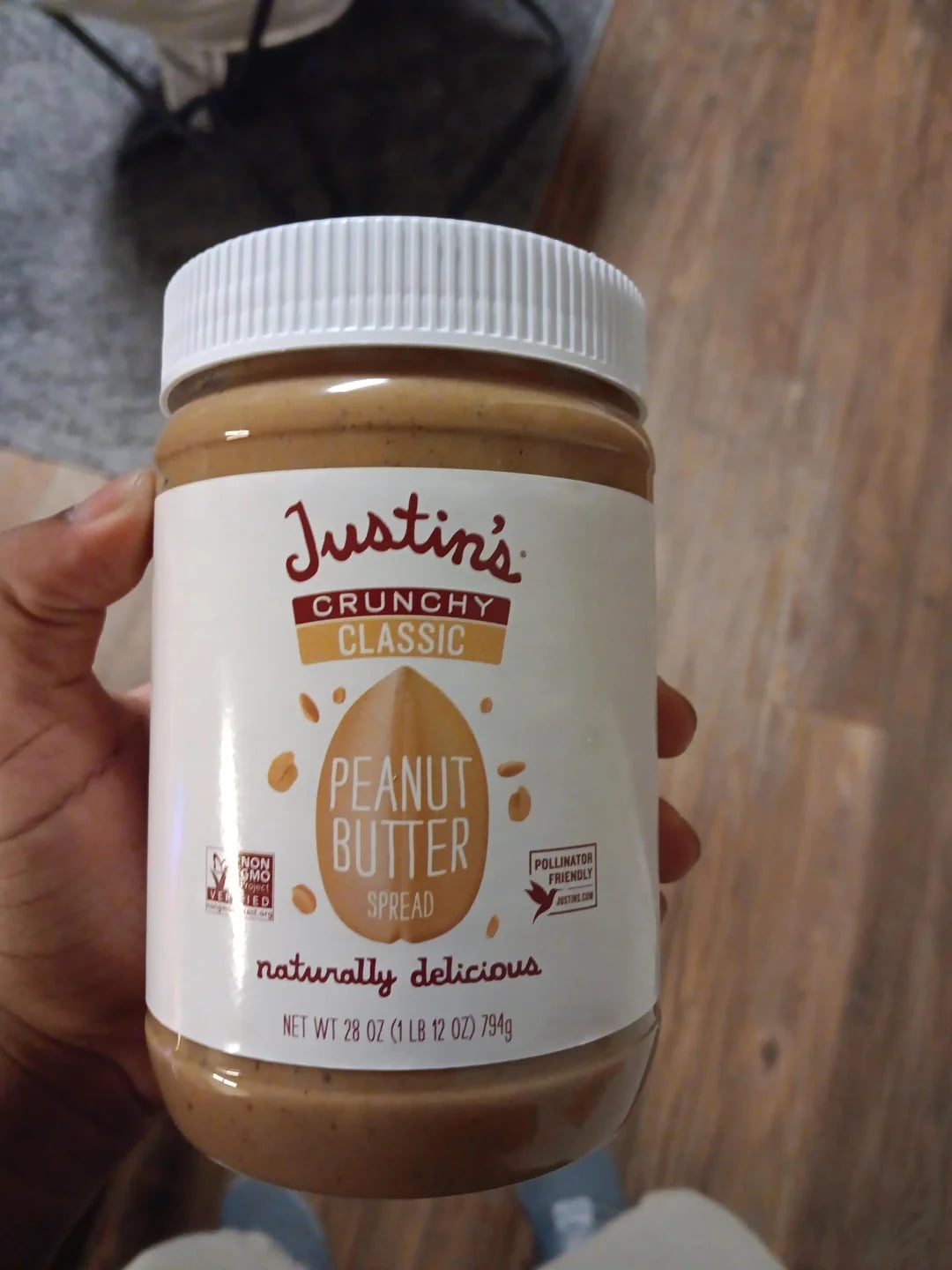 A hand holds a 28-ounce jar of Justin's Crunchy Classic Peanut Butter Spread, one of the best peanut butter brands, with a white lid and a label featuring brown and yellow text, set against a wooden floor background.