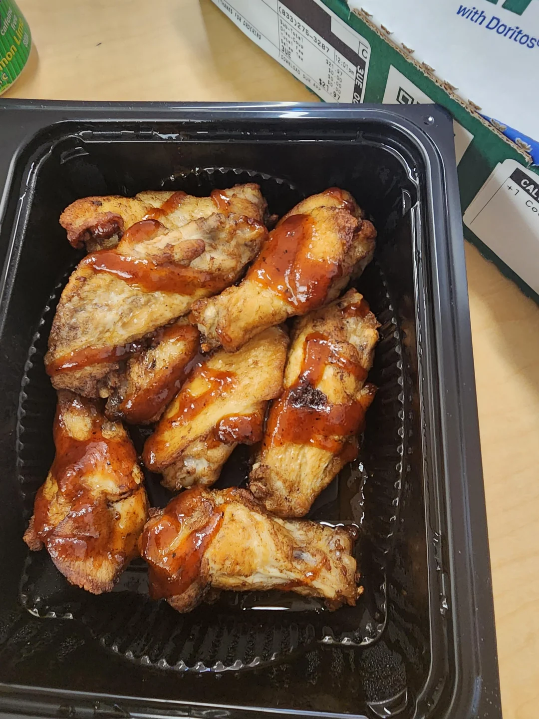 A black plastic container filled with cooked chicken wings drizzled with barbecue sauce, placed on a light-colored table. Part of a cardboard box and a can are visible in the background.