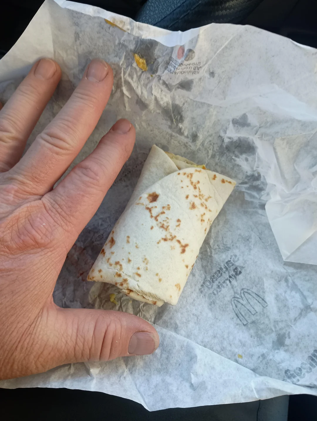 A hand rests next to a small wrapped tortilla breakfast burrito on crumpled McDonald's branded paper, showing the burrito's relatively small size.