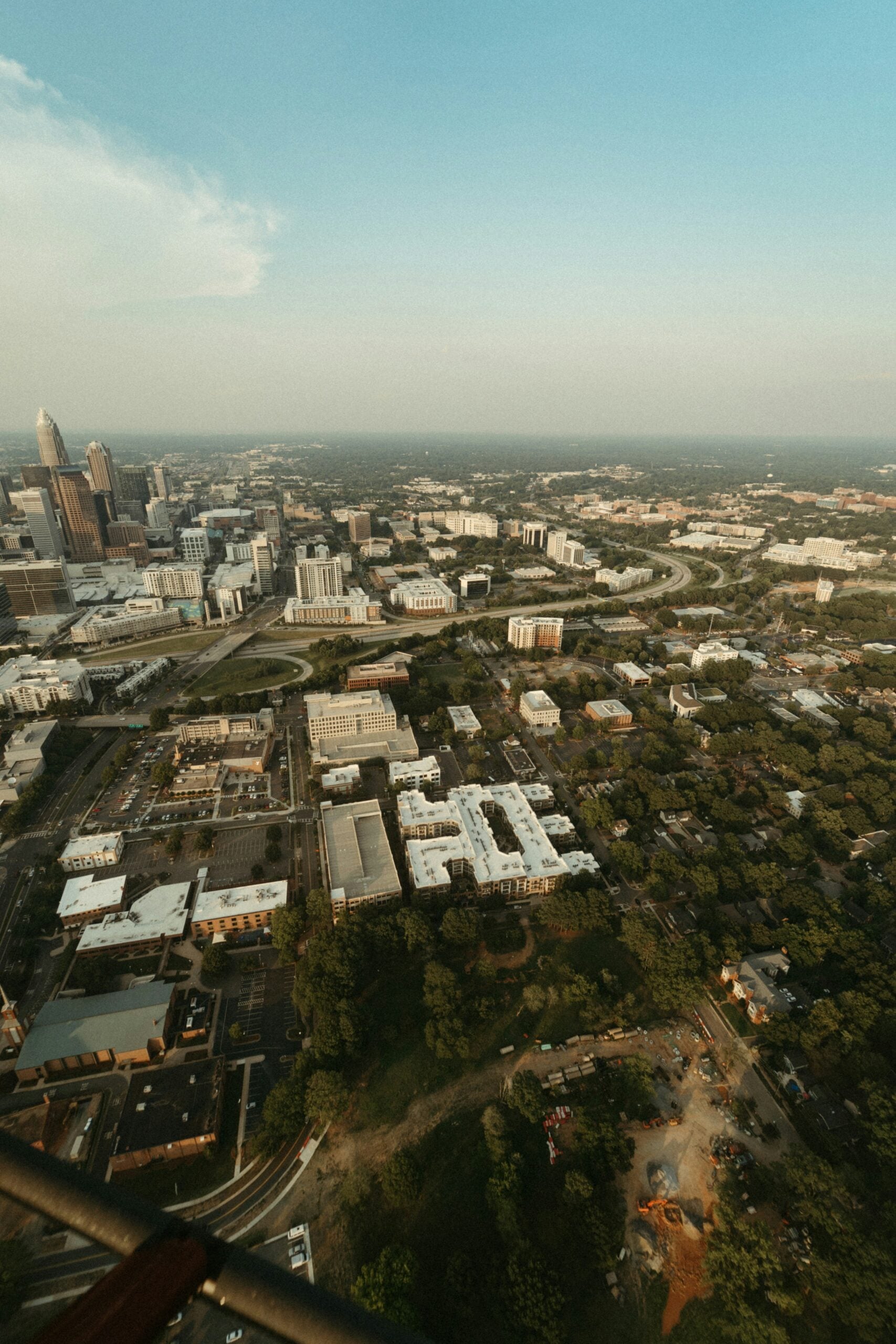 Aerial view of a city with tall buildings on the left, winding roads, clusters of white-roofed buildings, and green trees. The sky is clear with a hint of clouds on the upper left.
