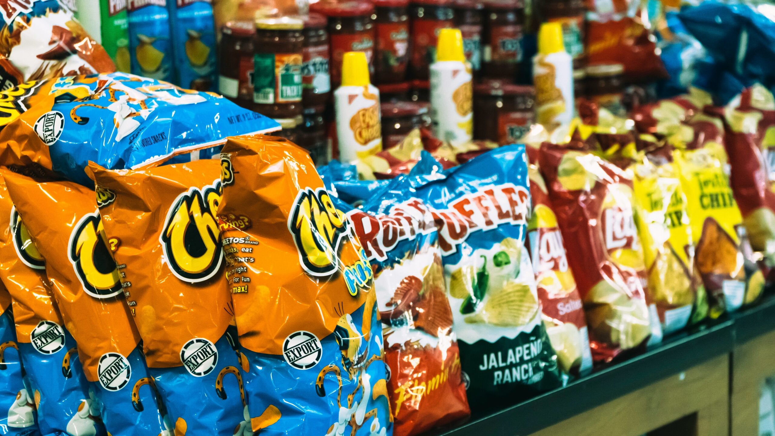 Bags of assorted chips, including Cheetos, Ruffles, and Lay’s, are displayed on a store shelf with jars of salsa and bottles of nacho cheese in the background.