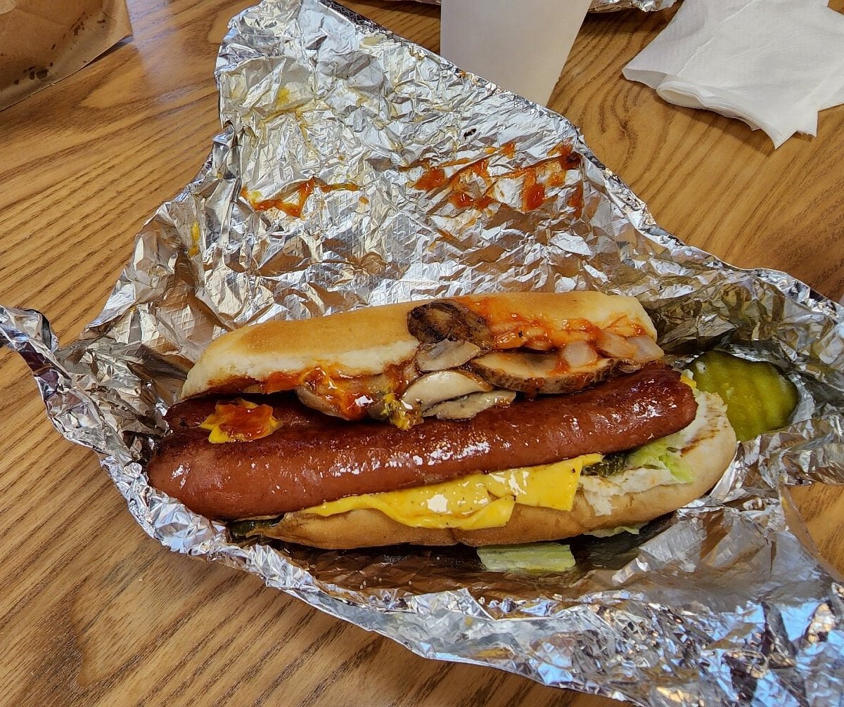 A Fast-Food Hot Dog with yellow cheese, lettuce, pickles, grilled mushrooms, and ketchup in a bun, partially wrapped in foil, sits on a wooden table next to a cup and napkins.