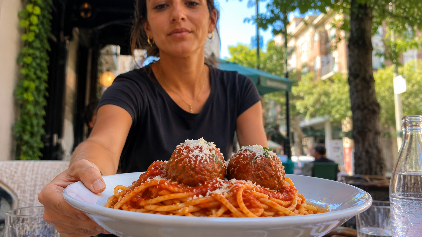 A woman at one of the best Italian restaurants sits outdoors, holding out a plate of spaghetti with tomato sauce and two large meatballs, topped with grated cheese. Trees and buildings are visible in the sunny background.