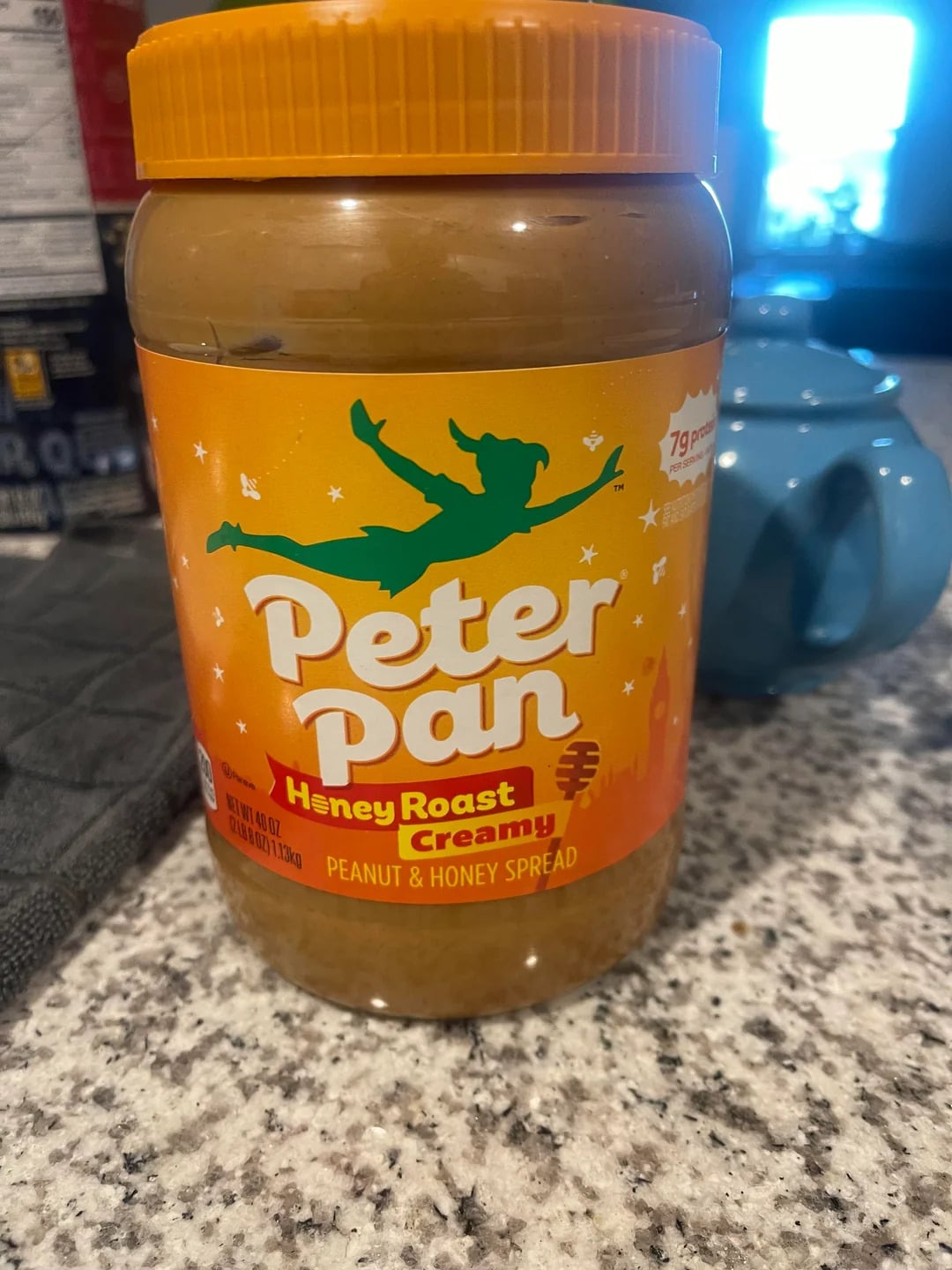 A jar of Peter Pan Honey Roast Creamy Peanut & Honey Spread, one of the best peanut butter brands, sits on a kitchen counter next to a blue teapot and a gray cloth. The orange lid and label feature a green silhouette of Peter Pan.