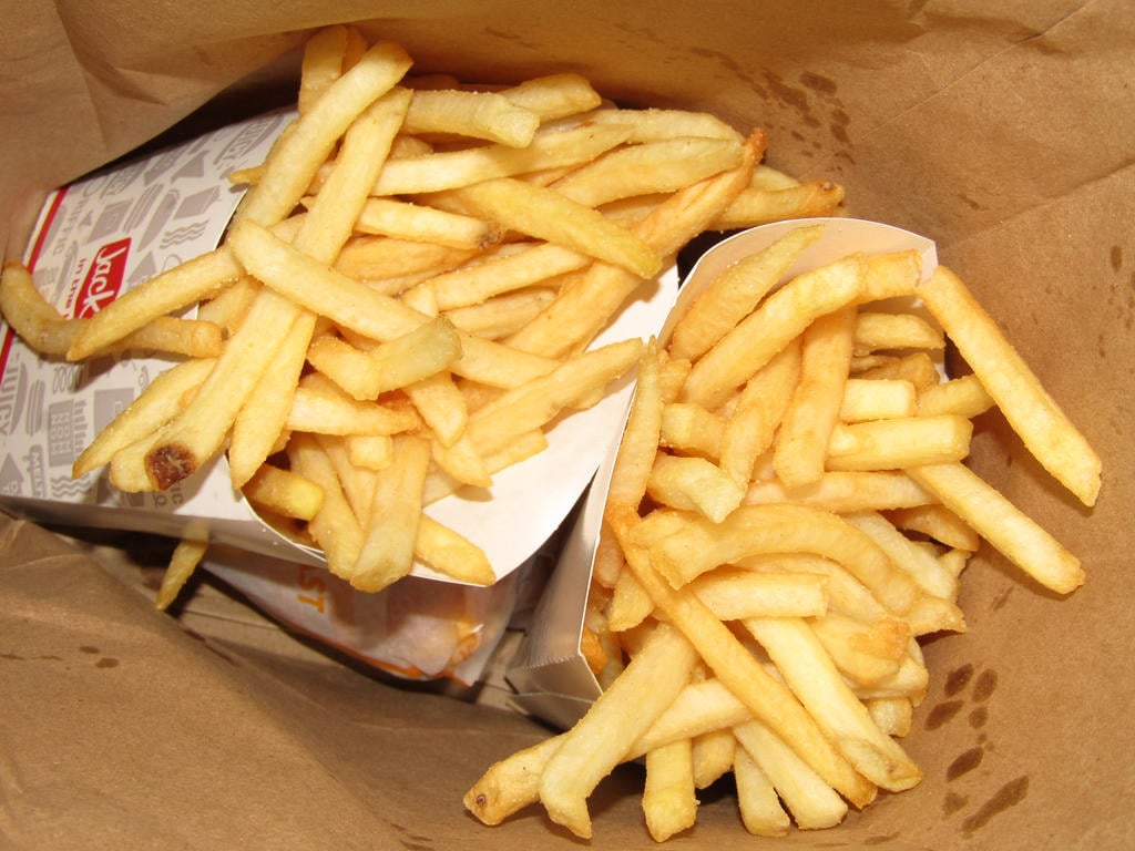 Two servings of French fries in paper containers inside a brown paper bag, viewed from above. The fries appear golden and freshly cooked.