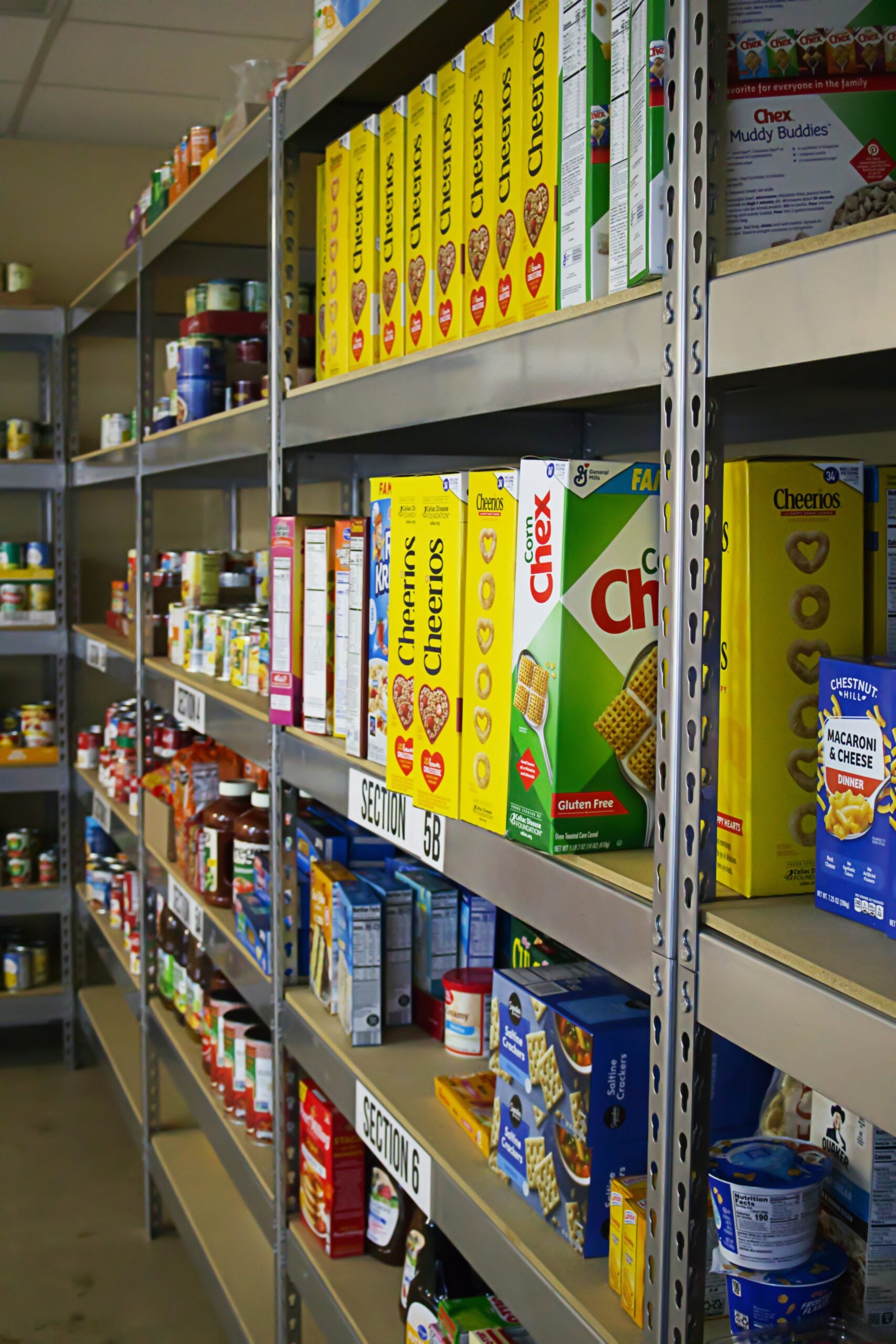 Shelves stocked with various food items, including cereal boxes like Cheerios and Chex, canned goods, and pasta, organized in sections inside a pantry or food bank.