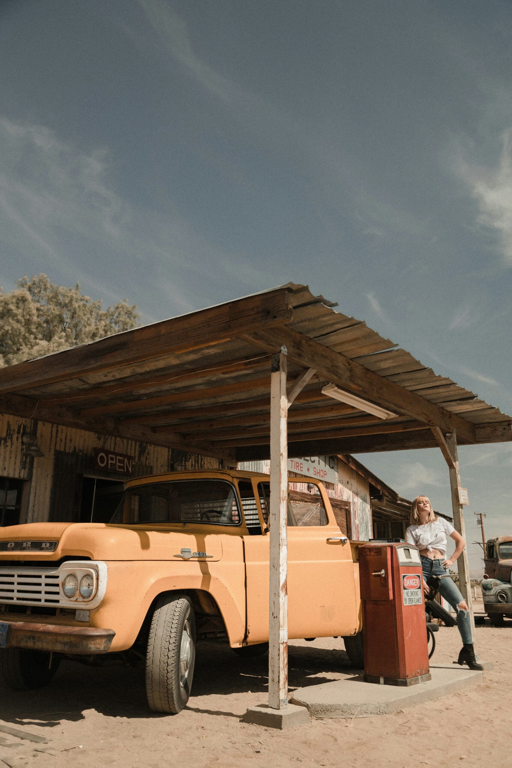 A person sits on the hood of a vintage yellow truck parked at an old-fashioned gas station with a wooden canopy, under a clear sky. The scene has a rustic, retro vibe.
