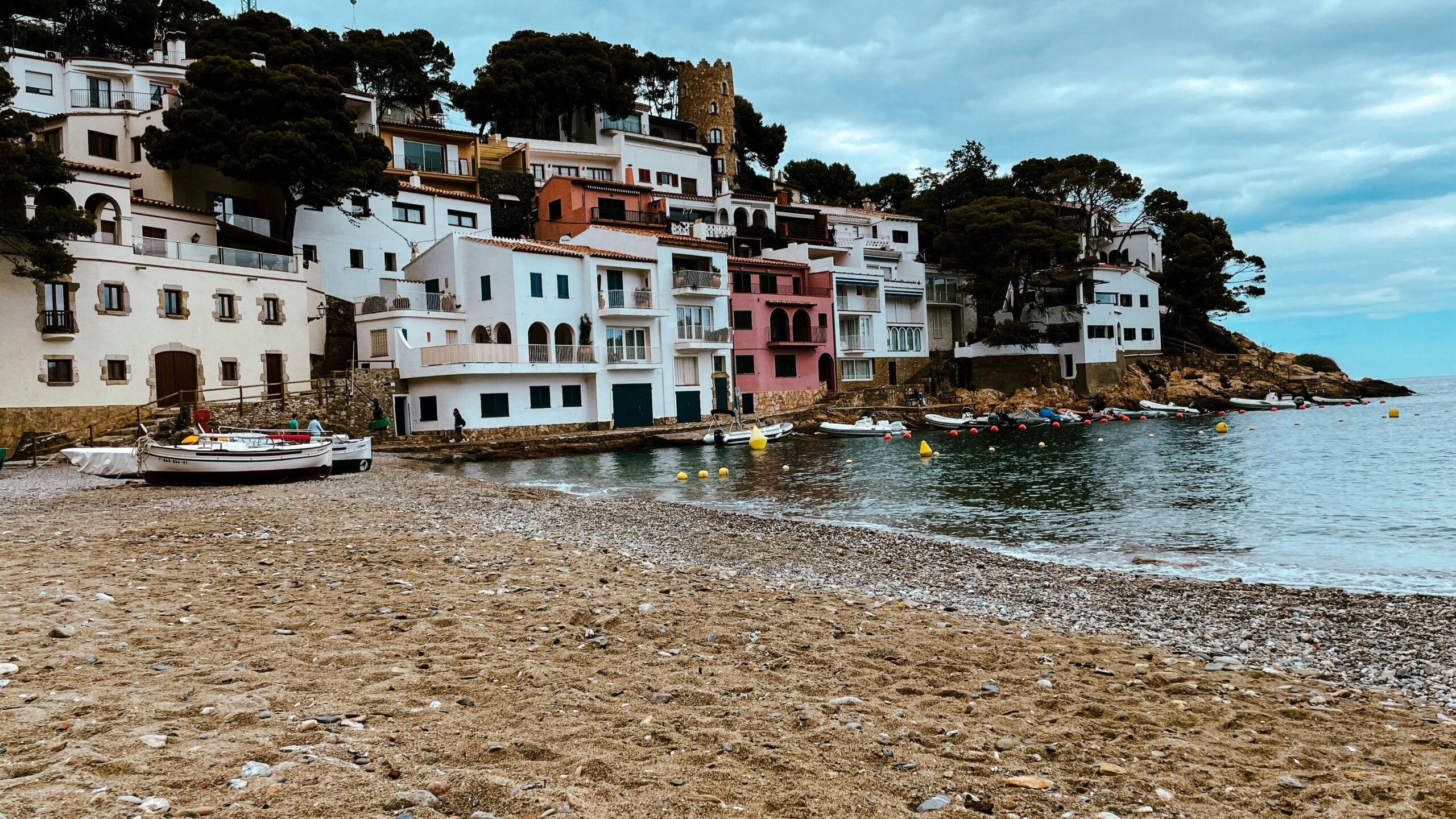 Colorful houses perched on a hillside overlook a small pebble beach with boats and calm water under a cloudy sky. Dense trees surround the buildings, and yellow buoys float near the shore.