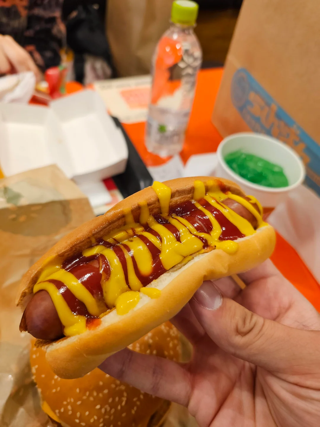 A hand holding a Fast-Food Hot Dog topped with ketchup and mustard; a sesame seed burger, green jelly dessert, bottled drink, and takeout containers are visible in the background on an orange table.