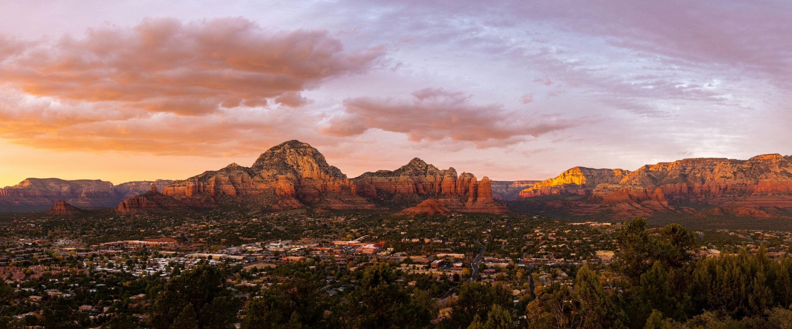 A panoramic view of Sedona, Arizona at sunset, showing red rock formations, scattered buildings, and trees in the foreground under a sky with scattered clouds tinted by the golden and pink hues of the setting sun.