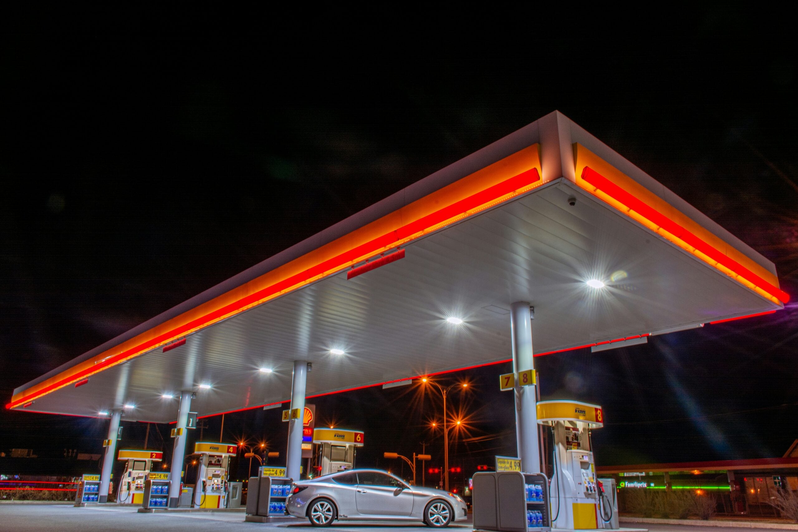Cheapest gas near major cities. A brightly lit gas station at night with a white car parked by the pumps. The canopy is illuminated with red and white lights, and the sky is dark in the background.