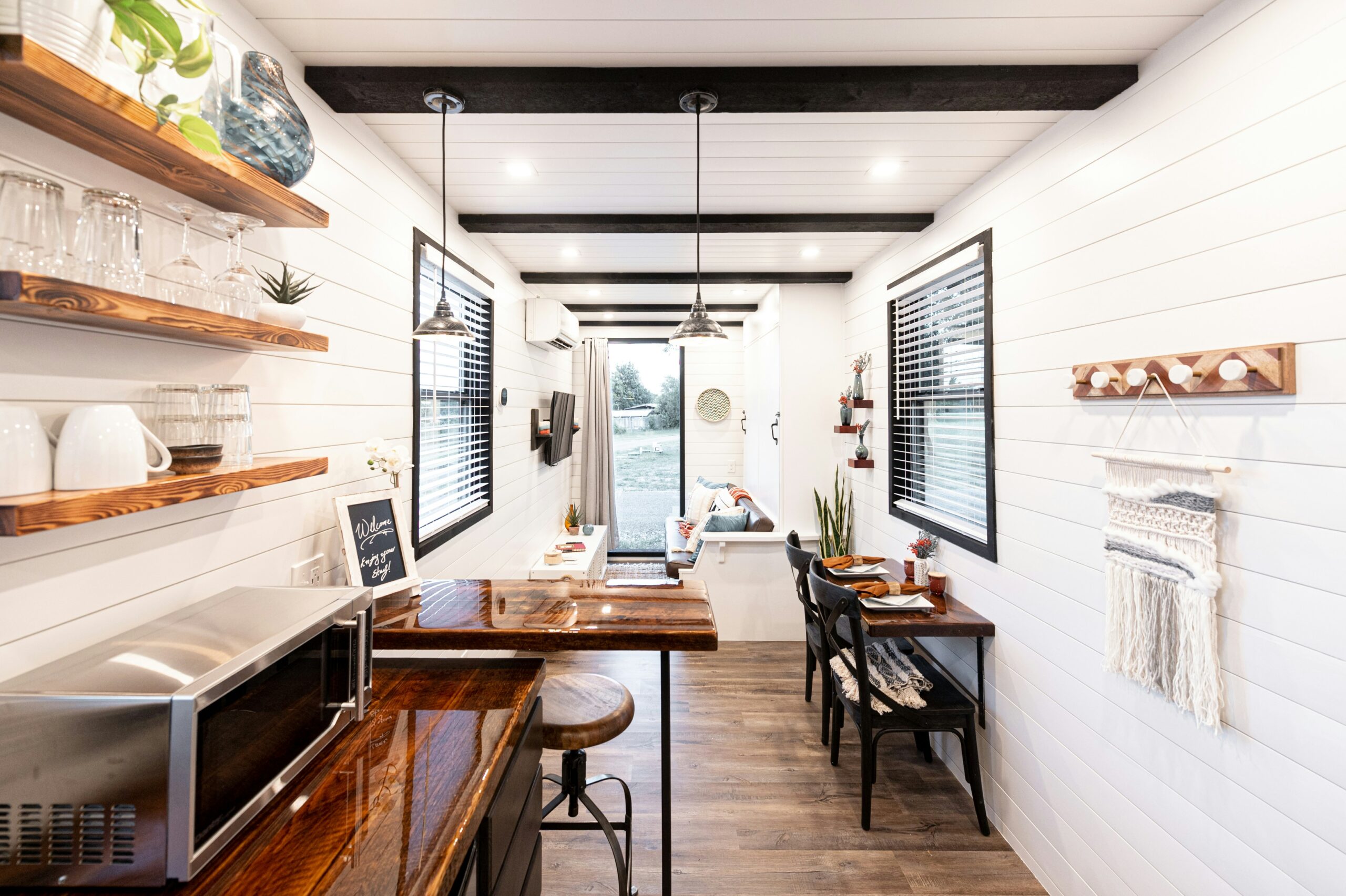 Bright, modern tiny house interior with white shiplap walls, wood accents, open shelving, a small kitchen with stools, and a cozy living area with large windows and natural light.