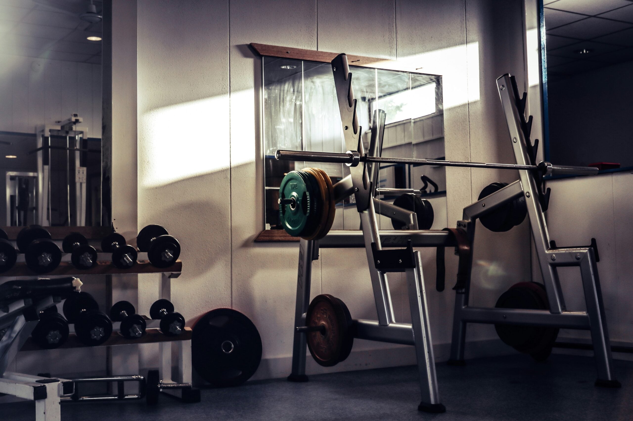 A gym with a barbell on a squat rack, weight plates loaded, and free weights neatly arranged on a rack nearby. Sunlight streams through a window, creating shadows on the floor and wall.