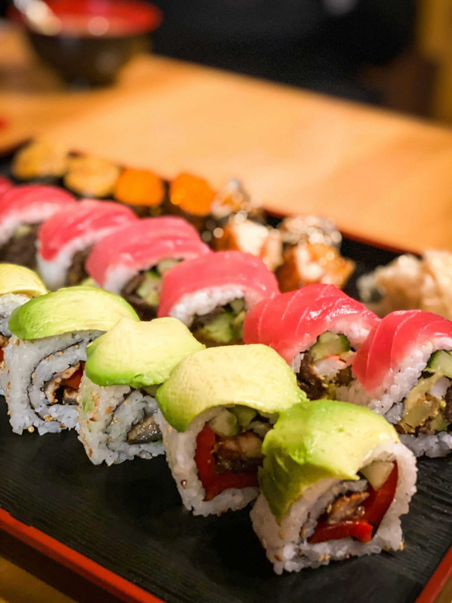 A close-up of assorted sushi rolls on a black tray, featuring rolls topped with avocado slices and tuna, with various colorful fillings visible inside the sushi.