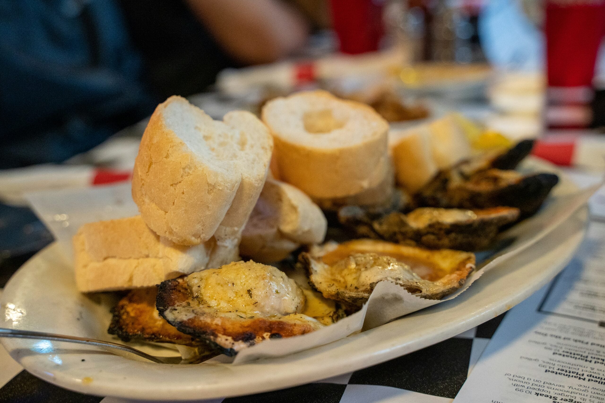 A plate of toasted oysters topped with cheese and herbs is served with thick slices of French bread on a checkered table, with red cups and menus in the background.