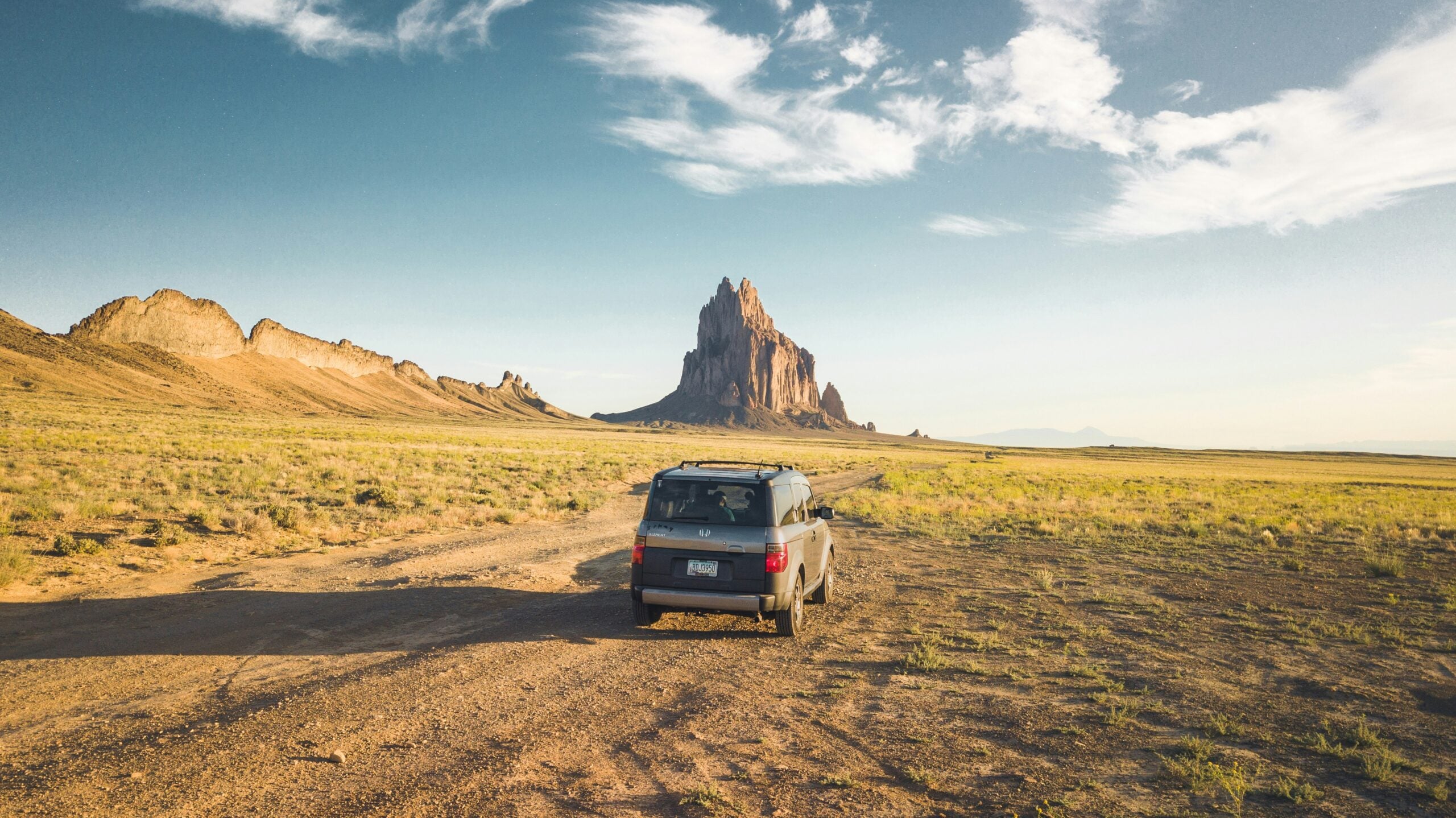 A car drives on a dirt road through a vast, open landscape with sparse grass, heading toward a tall, jagged rock formation under a partly cloudy blue sky.
