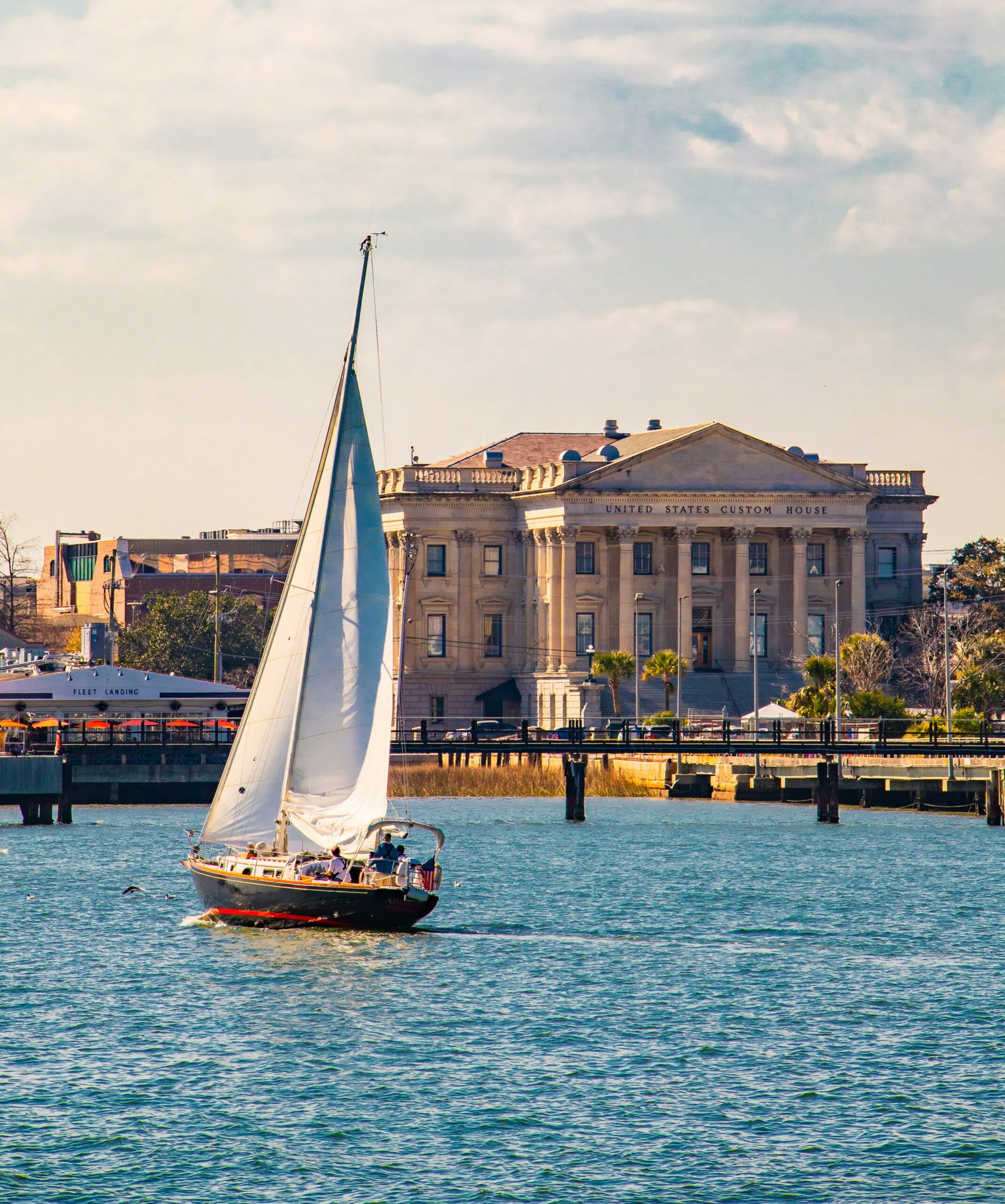 A white sailboat glides on calm blue water in front of the historic U.S. Custom House building under a partly cloudy sky. Trees and other buildings are visible in the background.