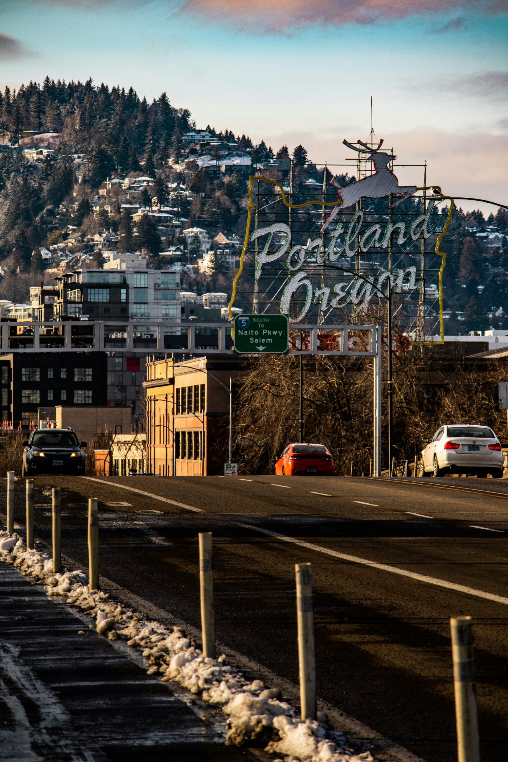 Cars drive toward the iconic “Portland Oregon” sign on a city street with snowy posts, urban buildings, and a forested hill in the background under a partly cloudy sky, capturing the spirit of the city as Cost of Living Relief measures take center stage.