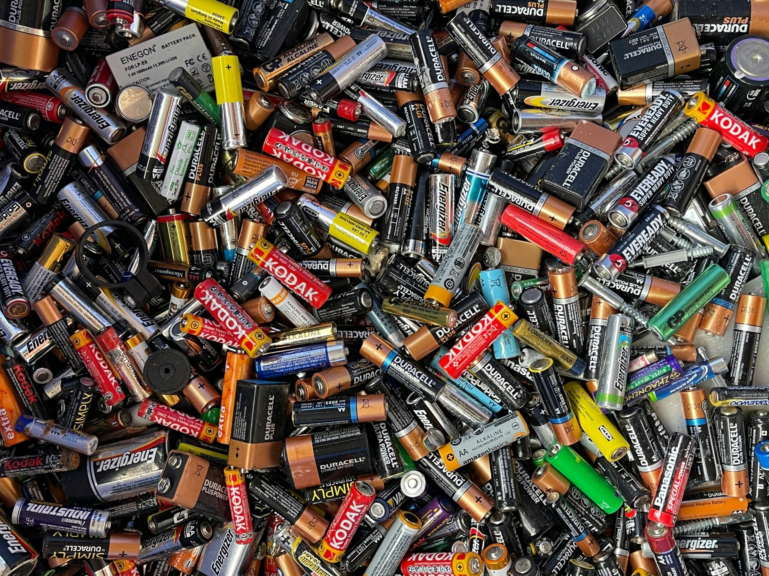 A large pile of assorted used batteries, including AA, AAA, C, D, and 9V sizes from various brands like Duracell, Kodak, and Energizer. The batteries are scattered randomly and appear worn.