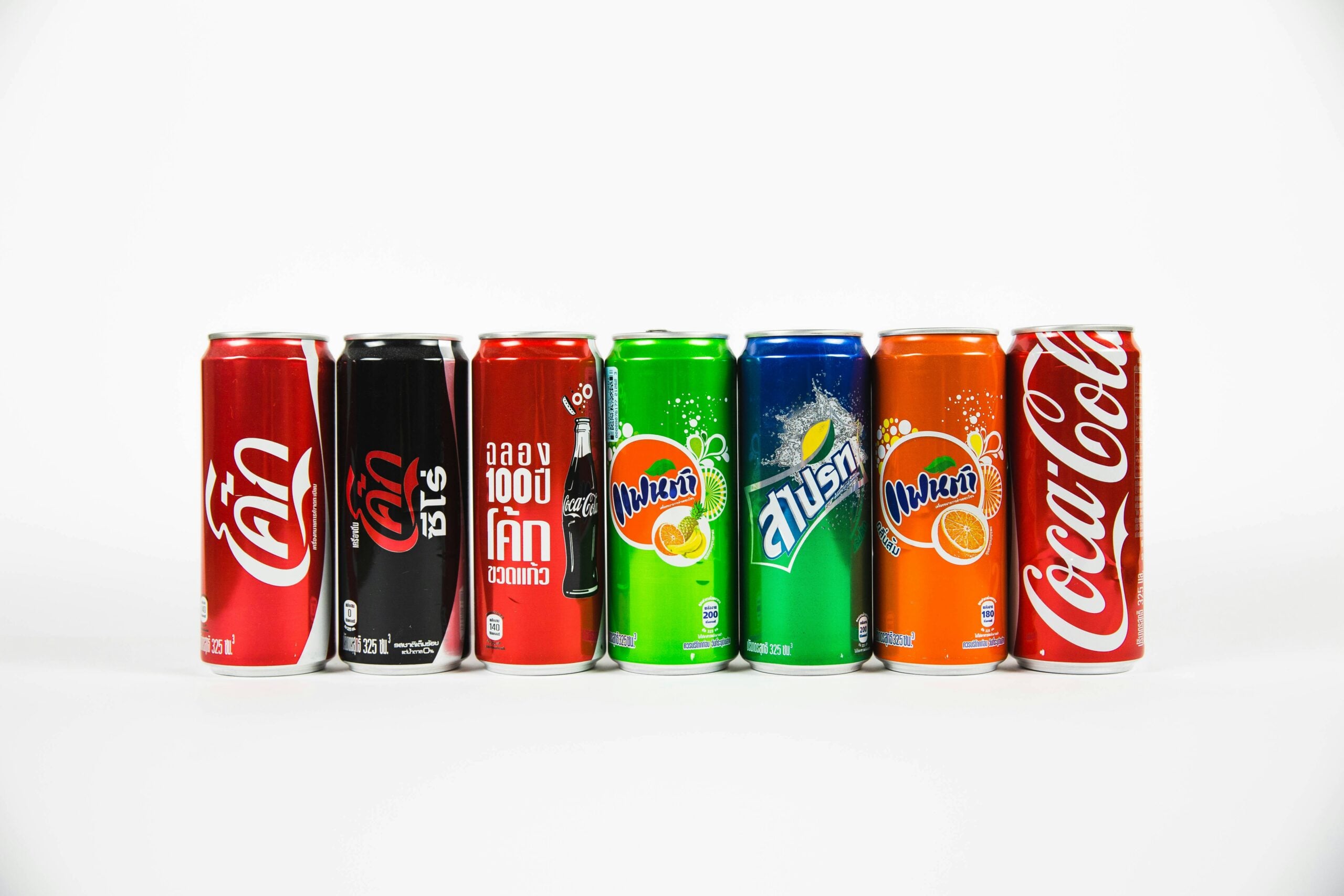 Seven colorful soda cans, including Coca-Cola, Coke Zero, and Fanta in various flavors, are lined up against a plain white background. The cans display both Thai and English text.