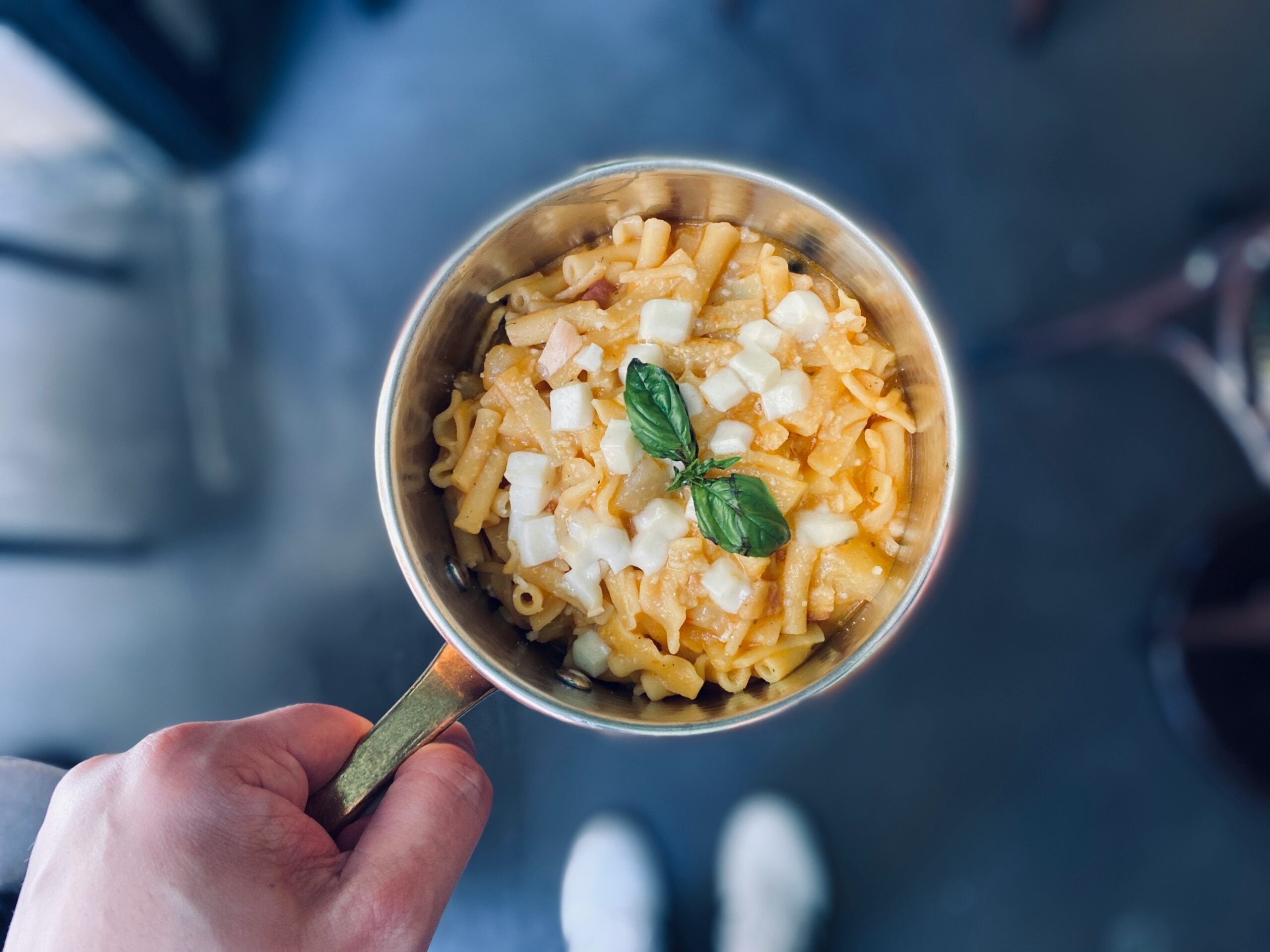 A hand holding a brass pot filled with macaroni and cheese, topped with chopped onions and fresh basil leaves. The background is blurred, showing a dark floor and the tips of white shoes.