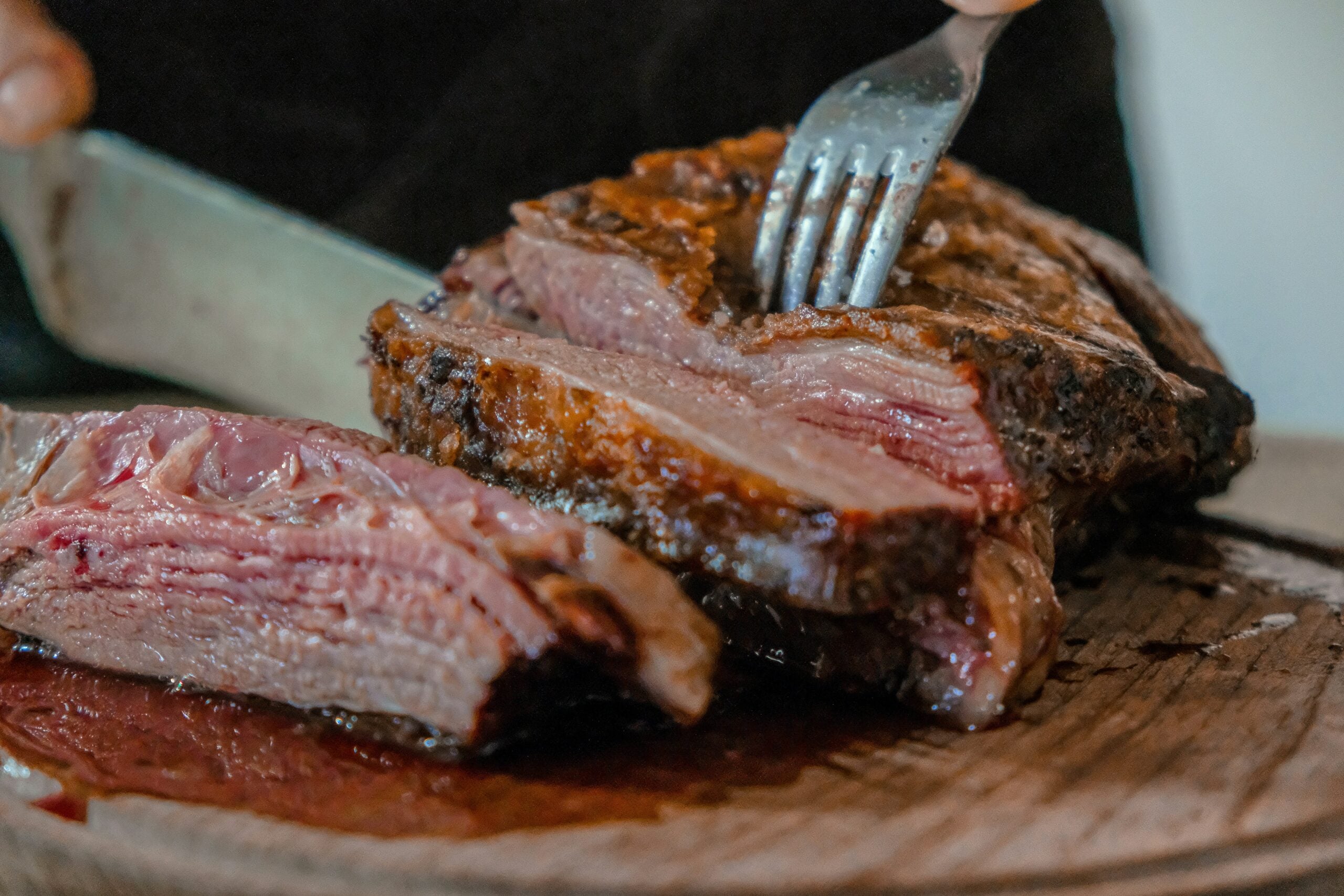 A close-up of a person slicing a juicy, medium-rare piece of roast beef on a wooden cutting board, with a fork holding the meat steady and visible juices on the surface.