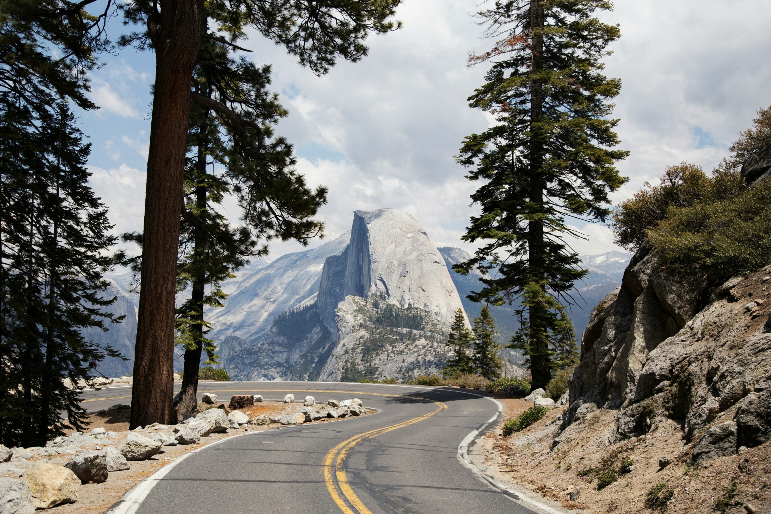 A winding mountain road curves between tall pine trees, leading toward the iconic granite peak of Half Dome under a partly cloudy sky in Yosemite National Park.