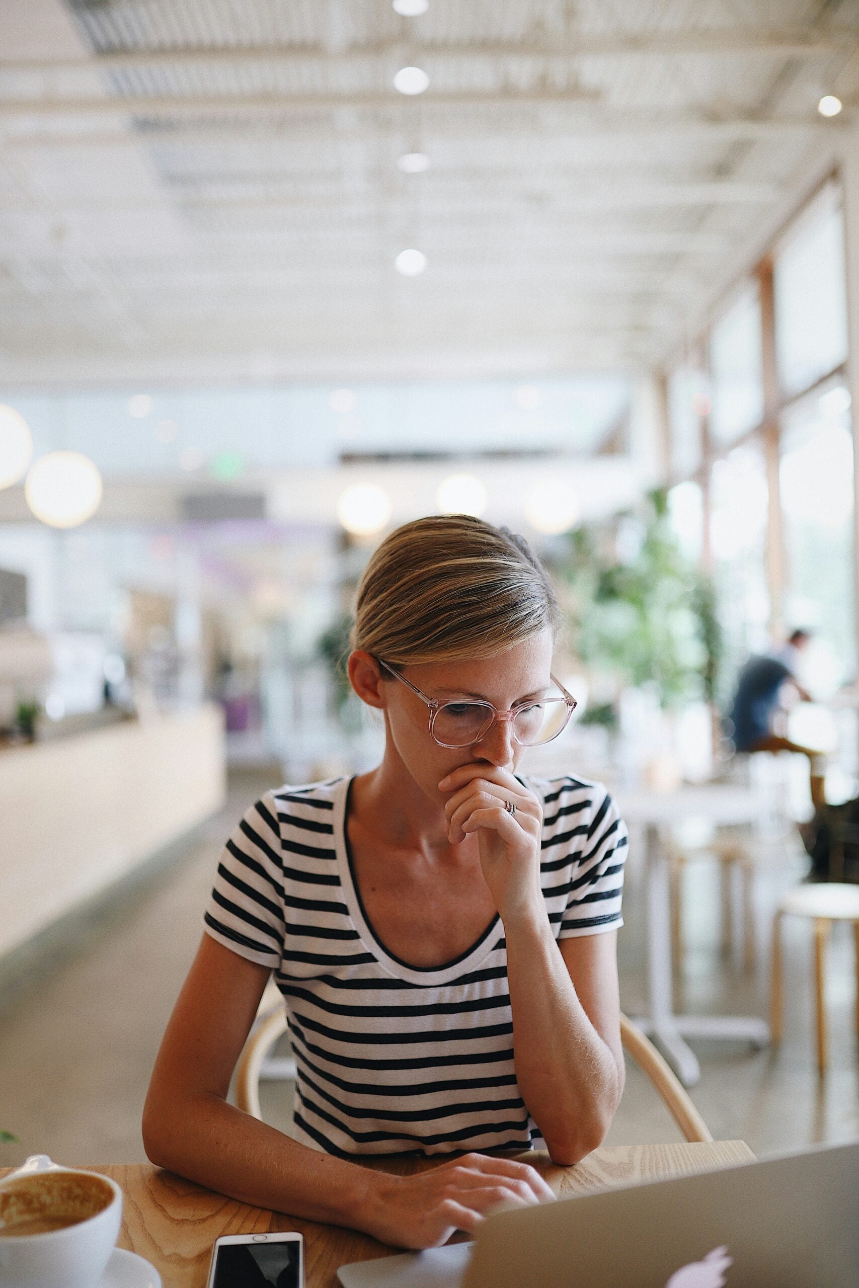 A woman wearing glasses and a striped shirt sits at a table in a bright cafe, looking thoughtfully at a laptop. A smartphone and a cup of coffee are on the table in front of her.