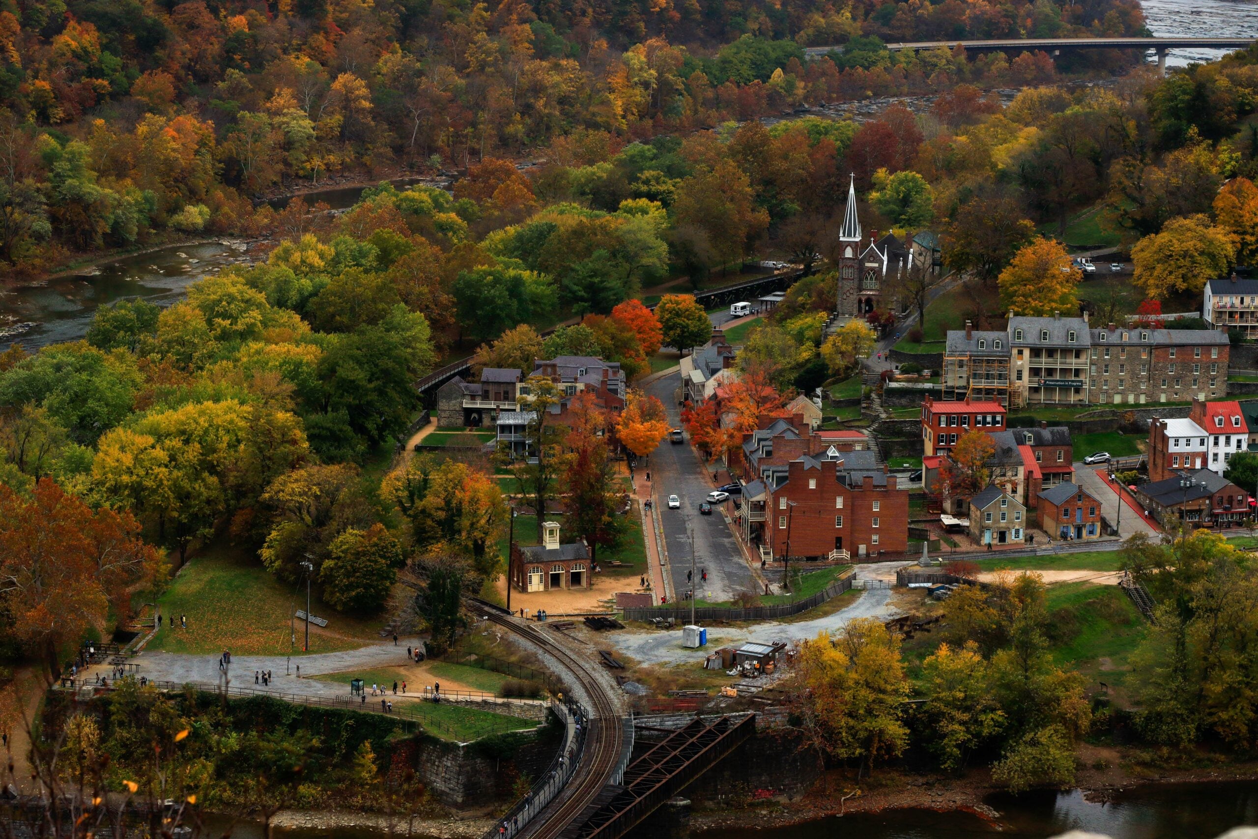 Aerial view of a small town surrounded by colorful autumn trees, with a river, a church, historic buildings, and a train track crossing over the water.