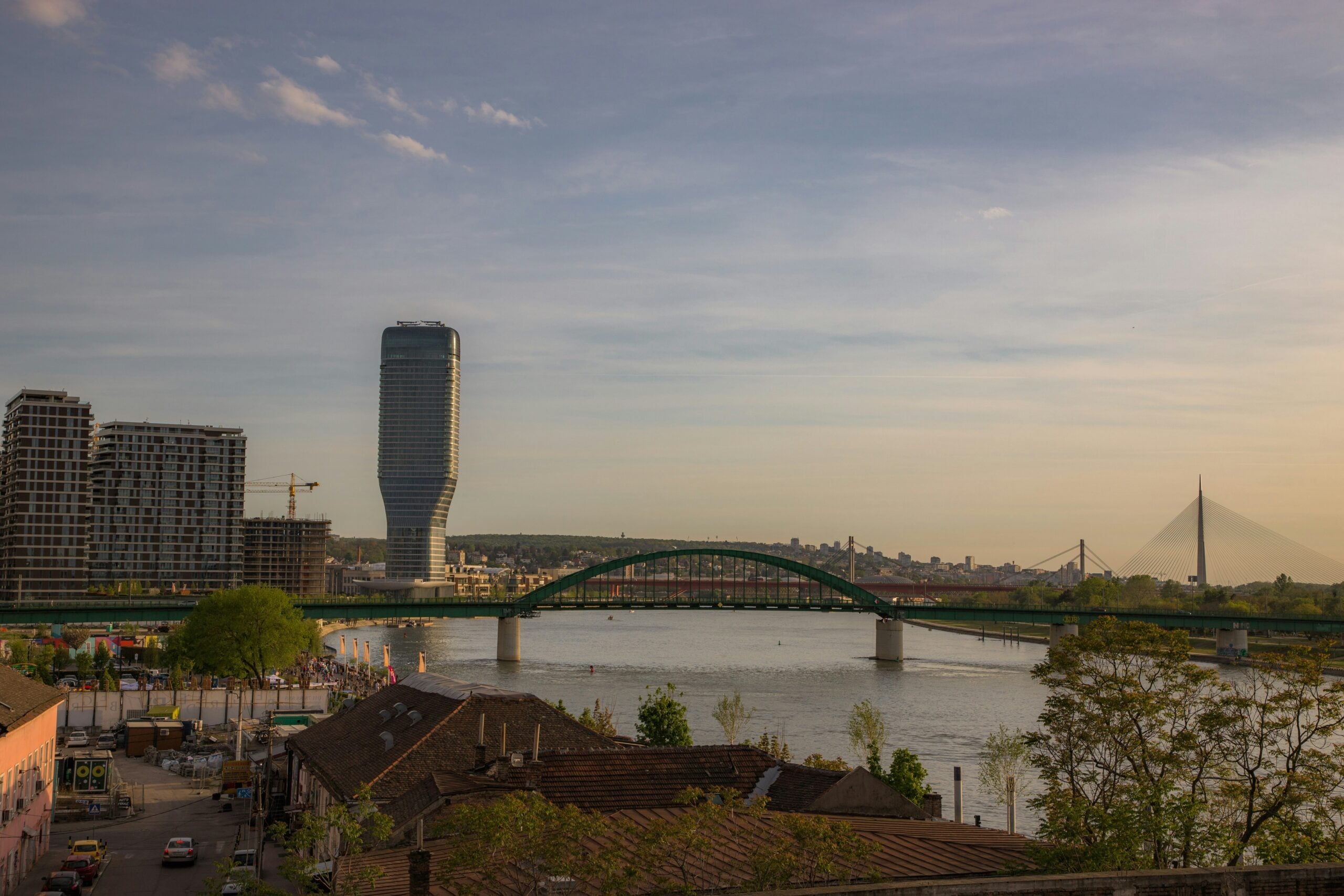 A view of a river with a green arch bridge, modern high-rise buildings on the left, and a cable-stayed bridge in the distance under a partly cloudy sky at sunset. Rooftops and trees are visible in the foreground.