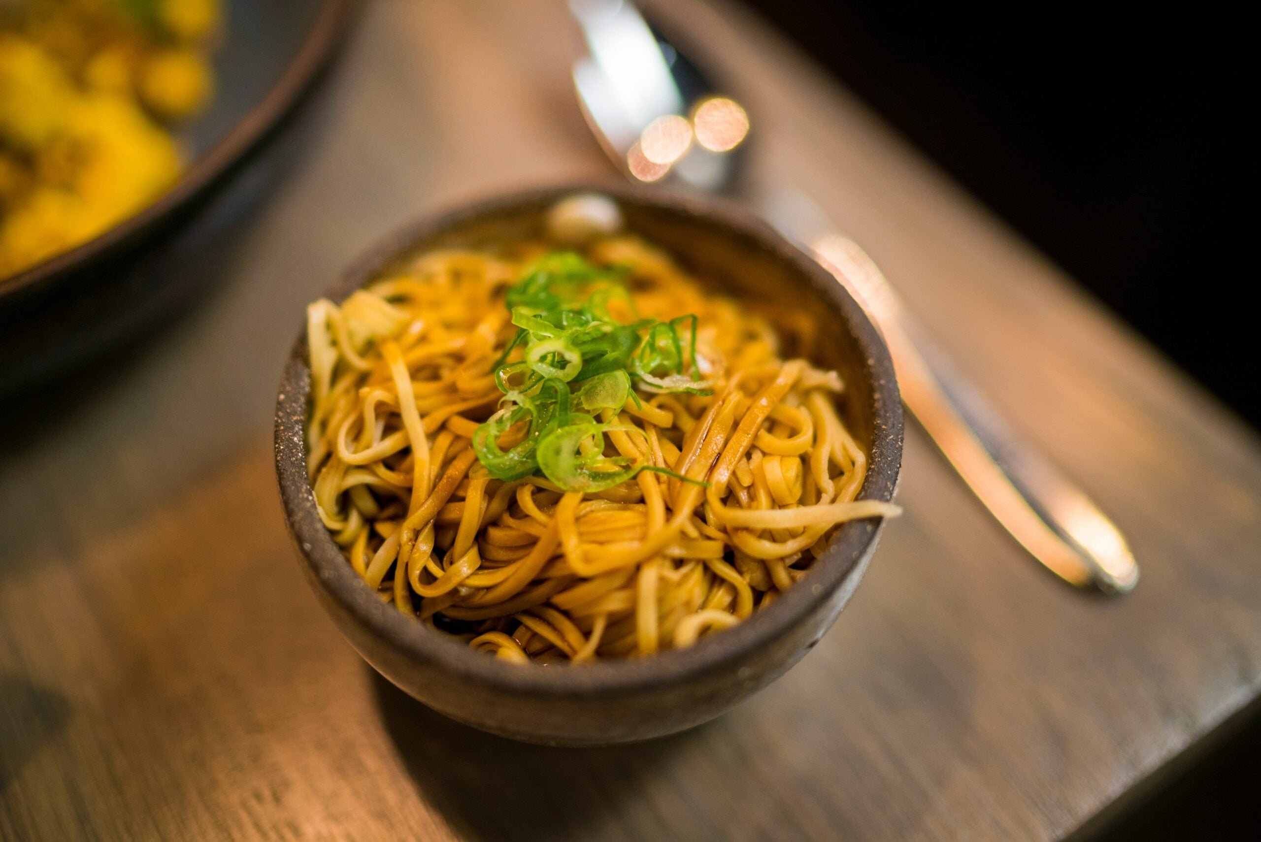 A bowl of cooked noodles garnished with sliced green onions sits on a wooden table, with a spoon and fork blurred in the background.