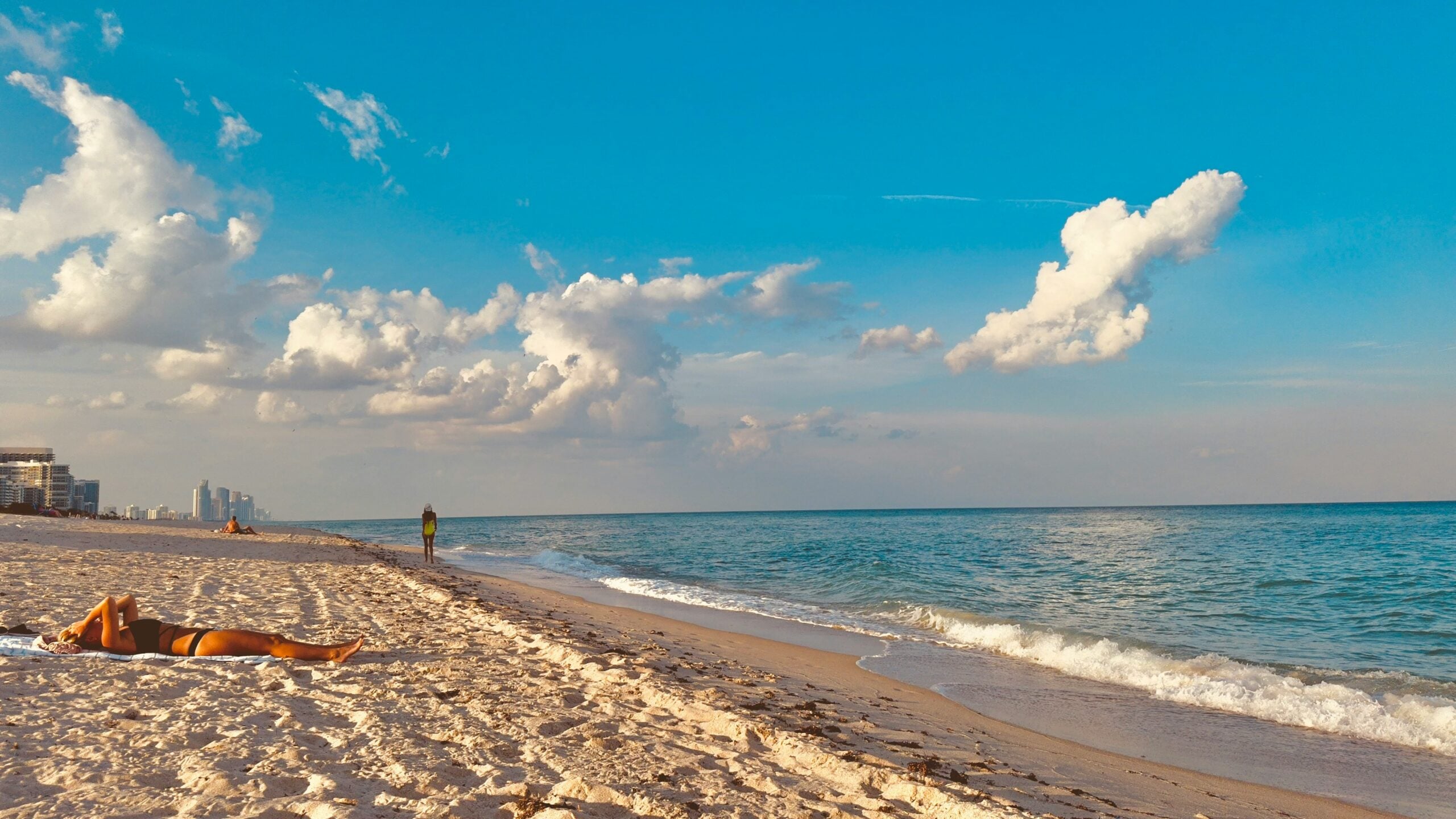 A person sunbathes on a sandy beach under a blue sky with scattered clouds; gentle waves roll onto the shore, and buildings are visible in the distant background.