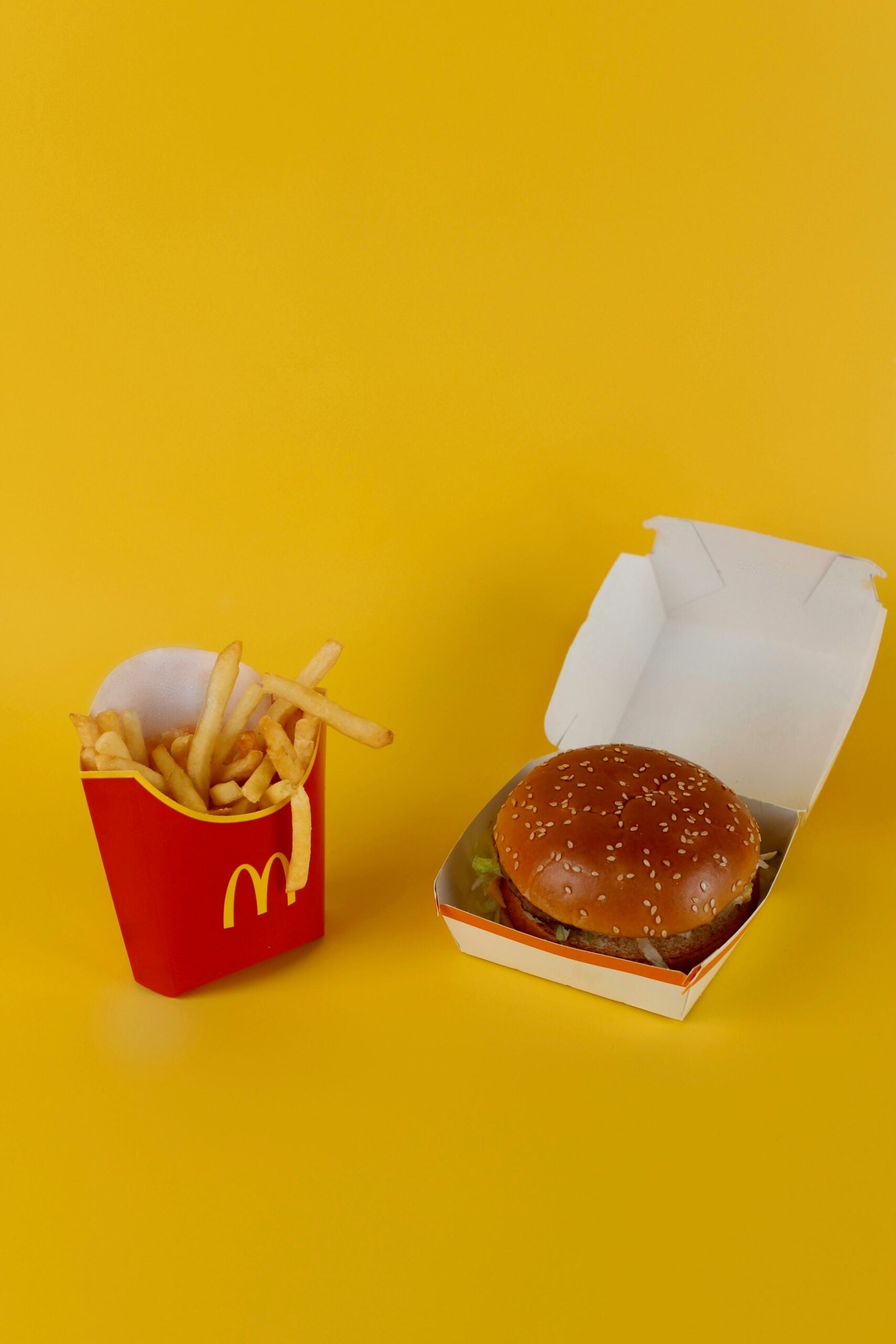 A McDonald’s medium fries in a red container and a sesame seed burger in an open white box are placed on a yellow background.