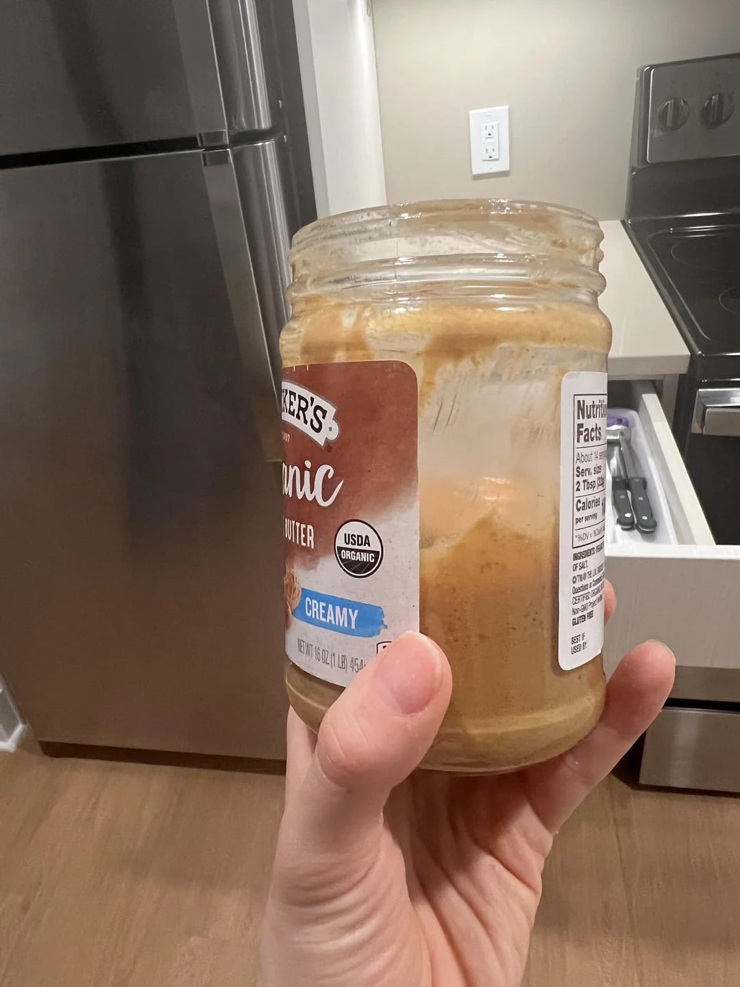 A hand holds a nearly empty glass jar from one of the best peanut butter brands in a kitchen, near a stainless steel refrigerator, stove, and light-colored cabinets.