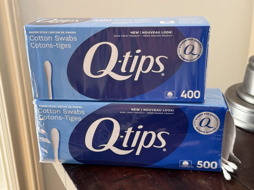 Two blue boxes of Q-tips cotton swabs, one box contains 400 swabs and the other 500. Both boxes are stacked on a dark wooden surface near a lamp.