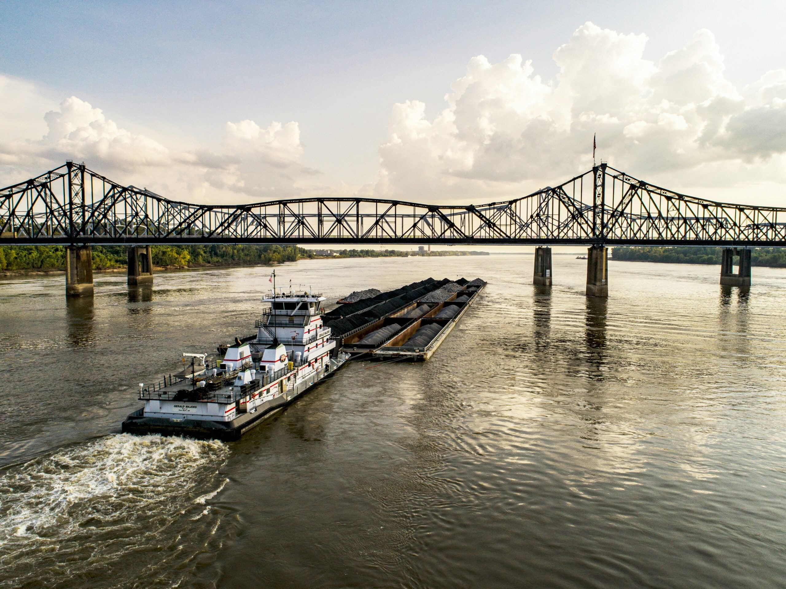 A barge carrying cargo travels down a river, approaching a large metal truss bridge under a partly cloudy sky, with trees visible along the distant riverbank.