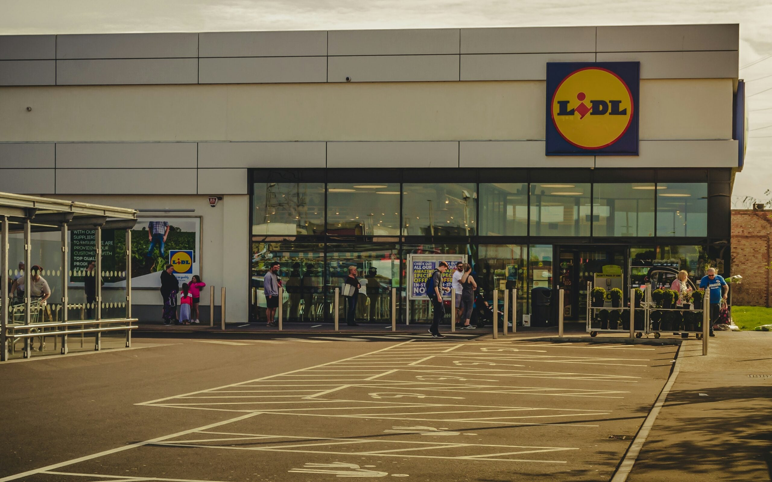 People are standing outside the entrance of a Lidl supermarket. Shopping carts are visible on the right, and there are parking spaces in front of the store. The Lidl logo is displayed above the entrance.