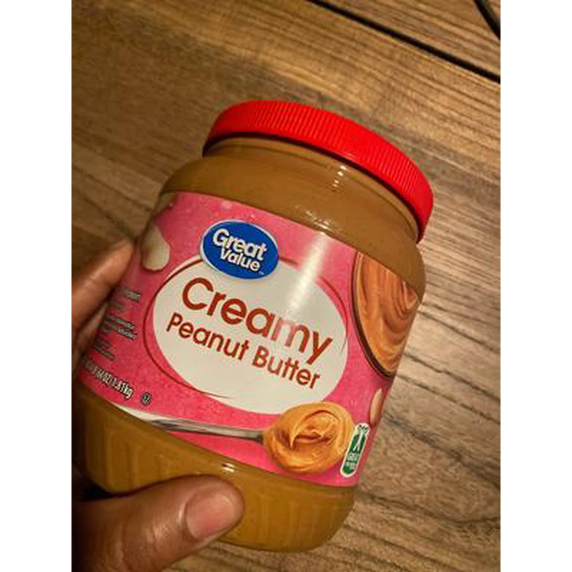 A hand holds a jar of Great Value Creamy Peanut Butter with a red lid, on a wooden surface. The mostly pink label features images of peanuts and a spoonful—one of the best peanut butter brands for creamy texture and classic flavor.