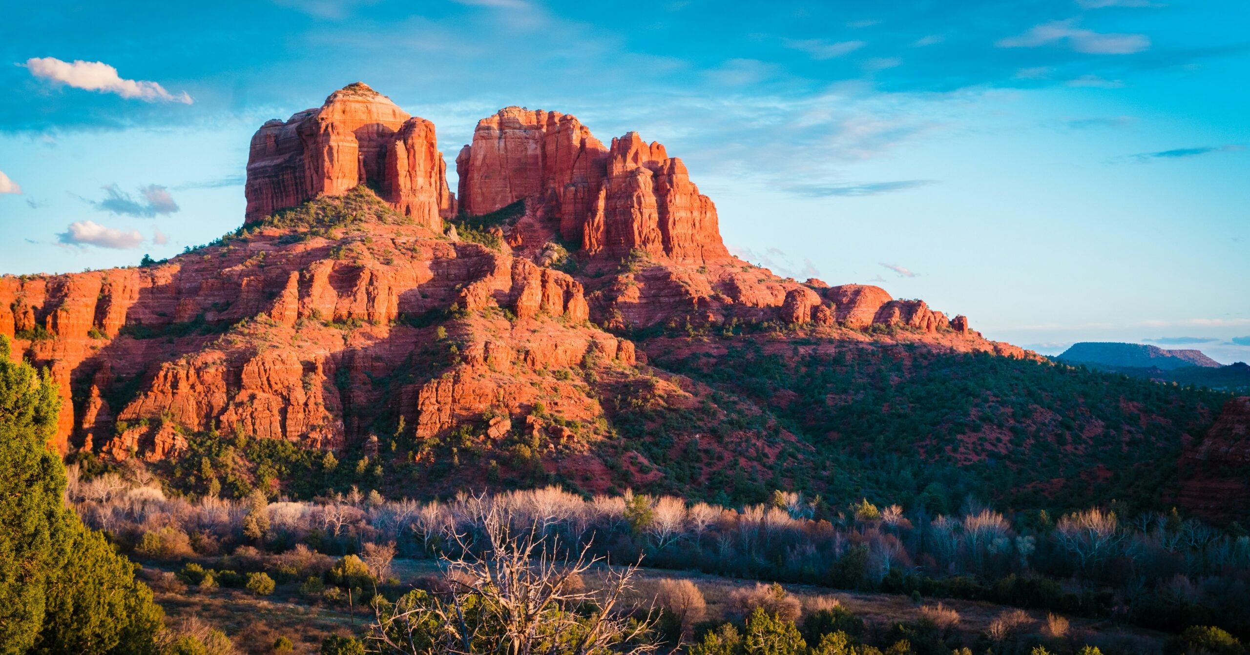 Large red rock formations rise above a green, brushy landscape under a blue sky with scattered clouds, illuminated by warm sunlight.