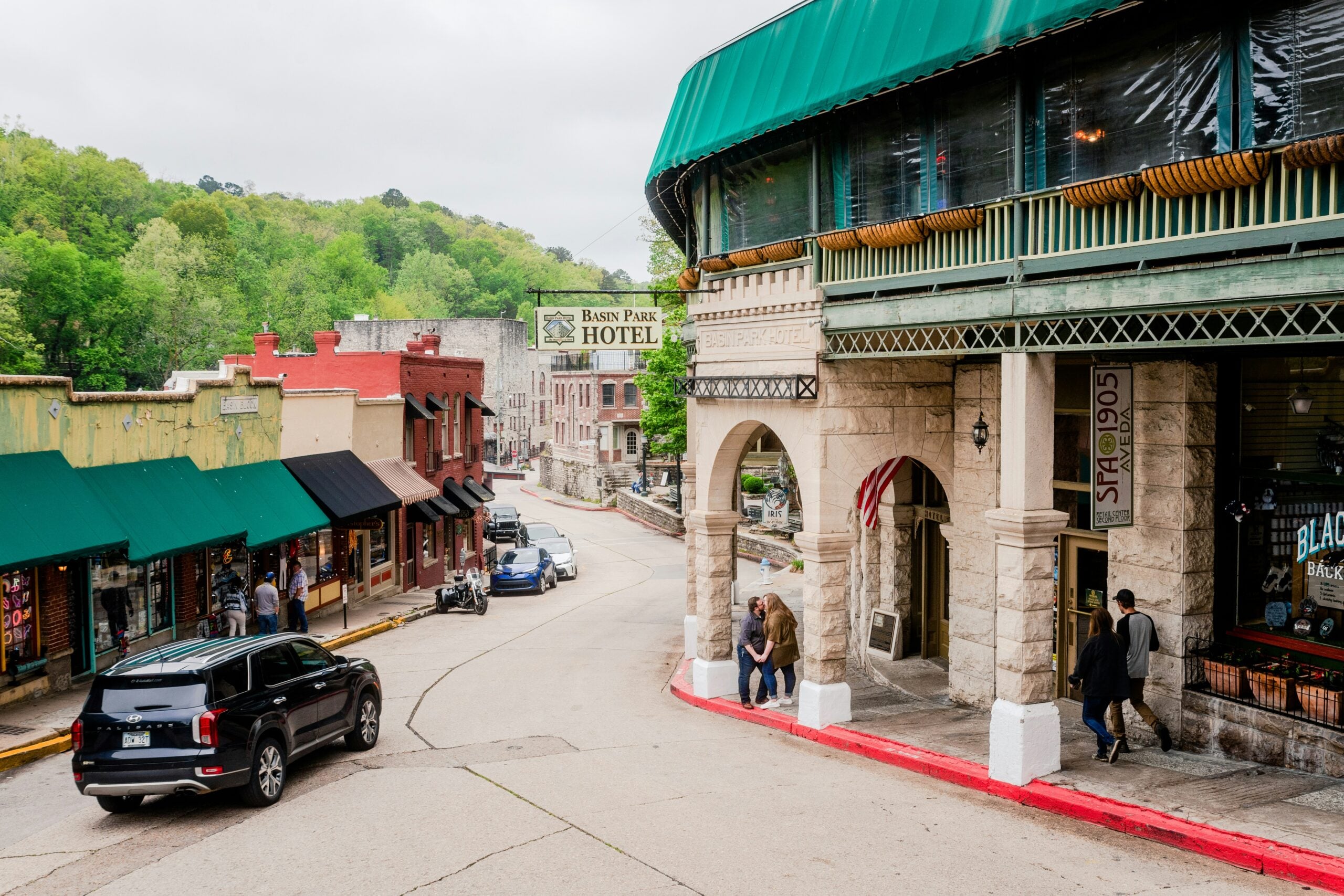 A curved street in one of the Unique American Towns, lined with historic buildings like the Basin Park Hotel, has parked cars, people strolling, and lush greenery in the background on a cloudy day.