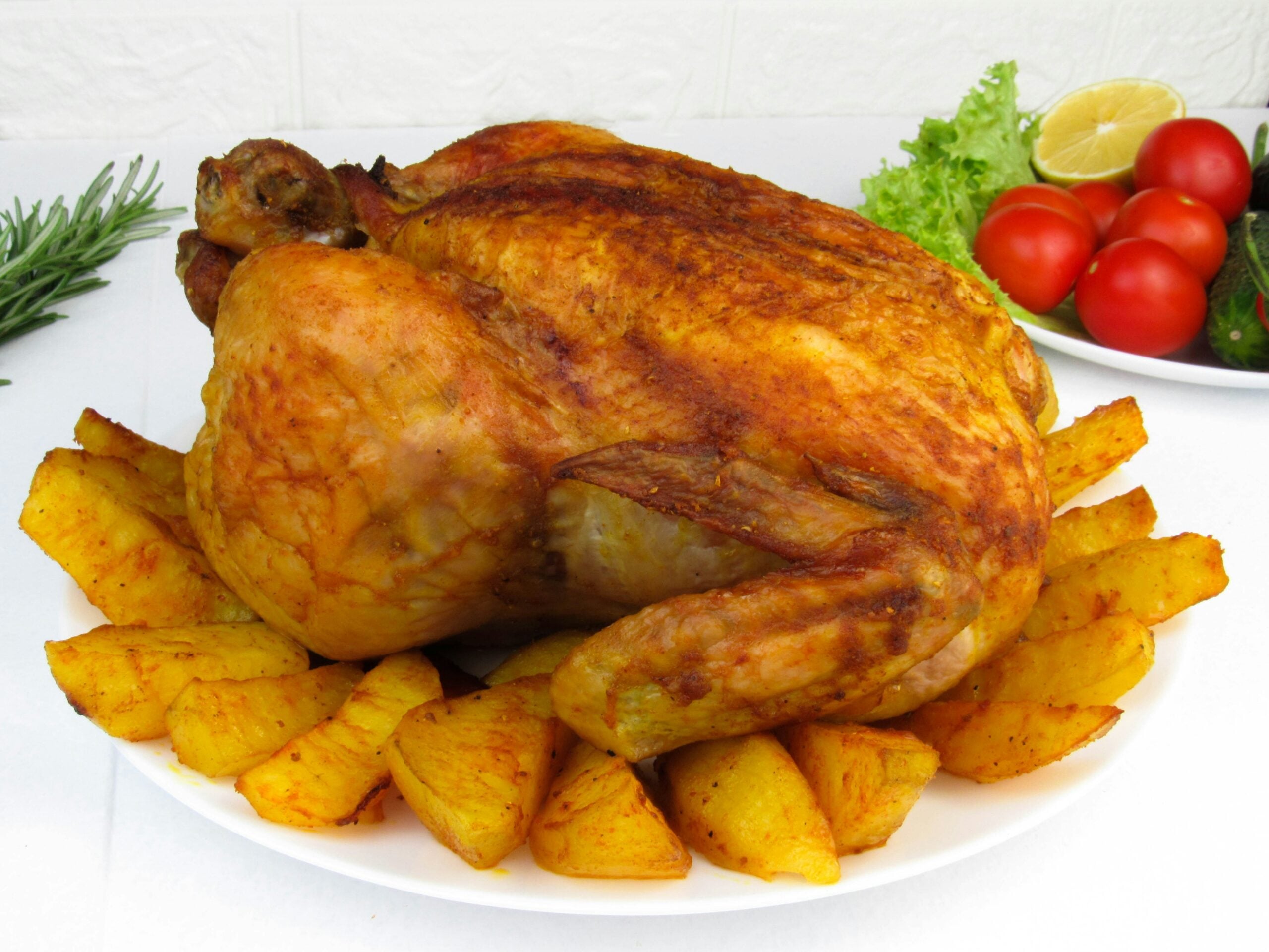 A whole roasted chicken served on a white plate surrounded by golden-brown potato wedges, with fresh vegetables like lettuce, tomatoes, cucumber, and lemon in the background.