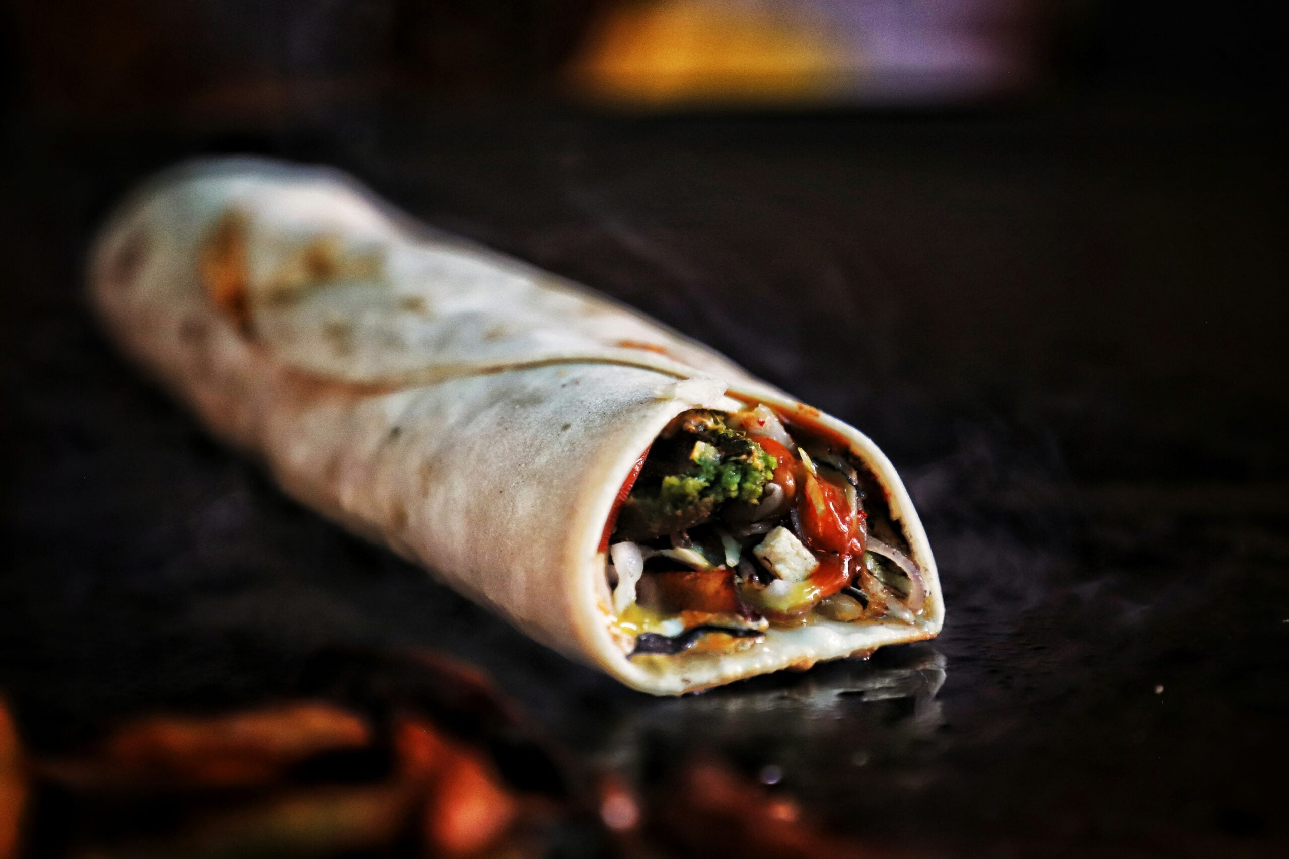 A close-up of a rolled tortilla wrap filled with colorful vegetables, placed on a dark surface with a blurred background.