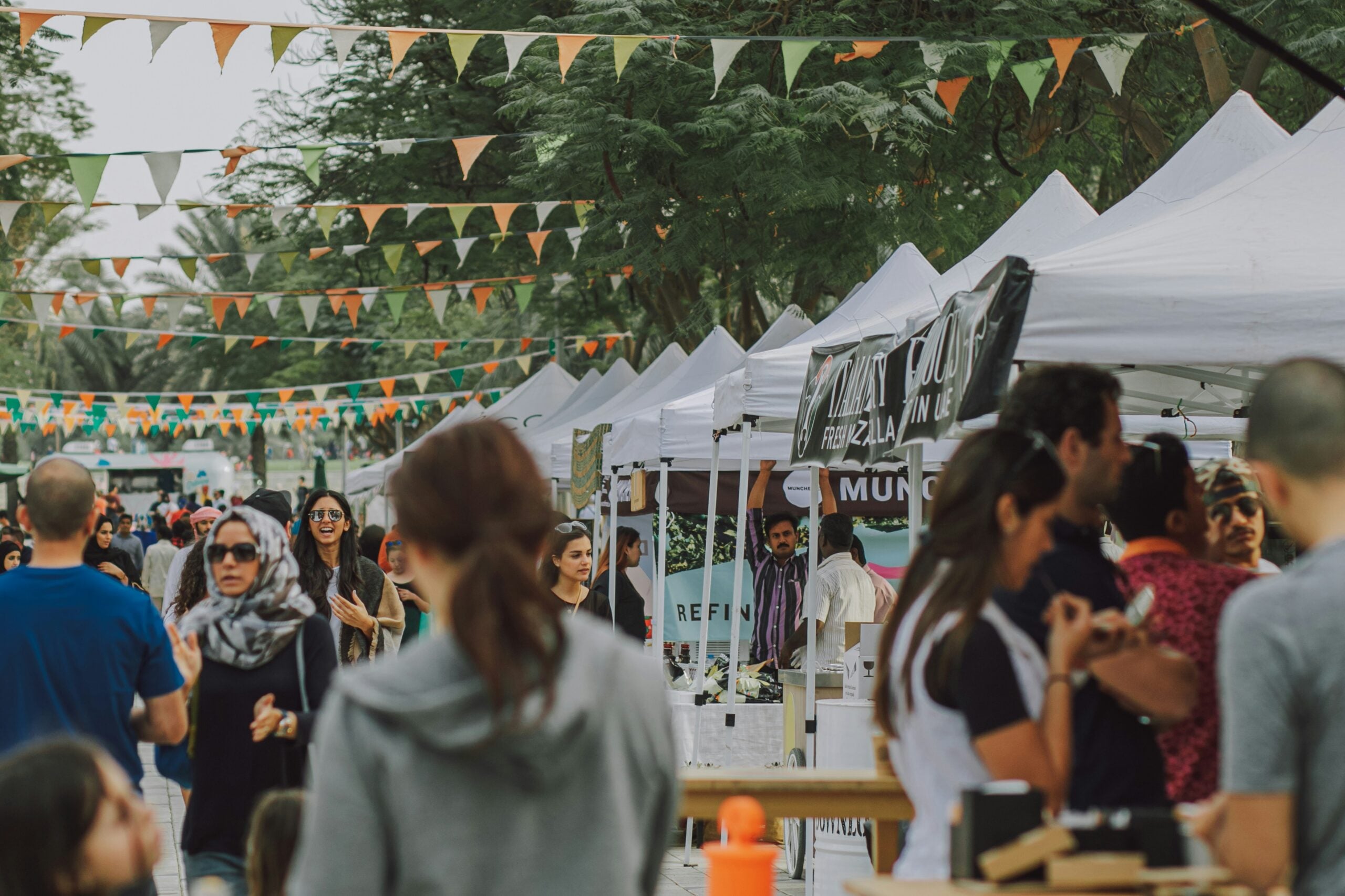 People walk and mingle at an outdoor market with white tents, colorful bunting flags overhead, and trees in the background. Various stalls line both sides, and the atmosphere appears lively and social.