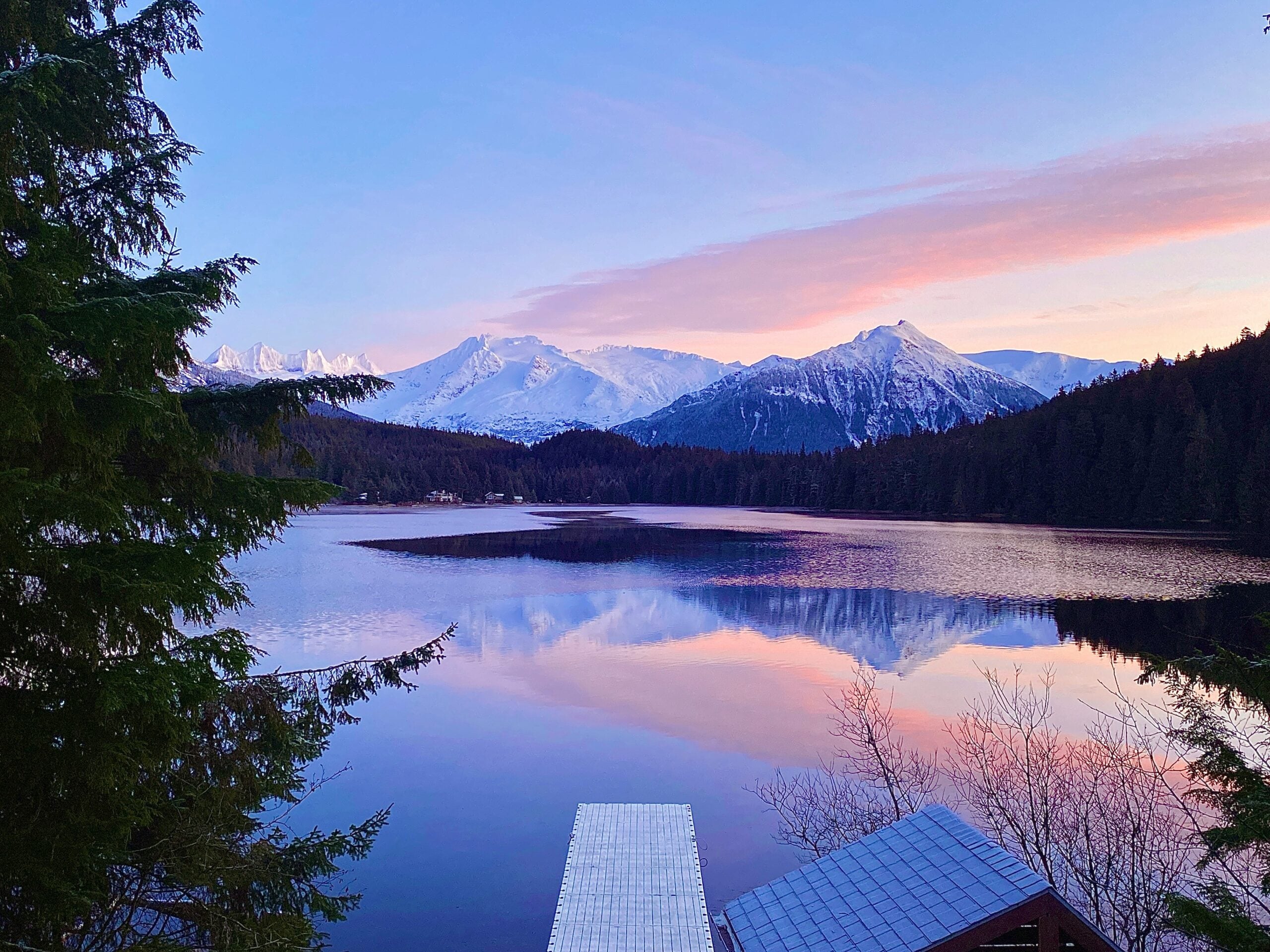 A serene lake reflects snow-capped mountains and a pink-blue sunset sky, surrounded by evergreen trees. A dock and part of a rooftop are visible in the foreground.