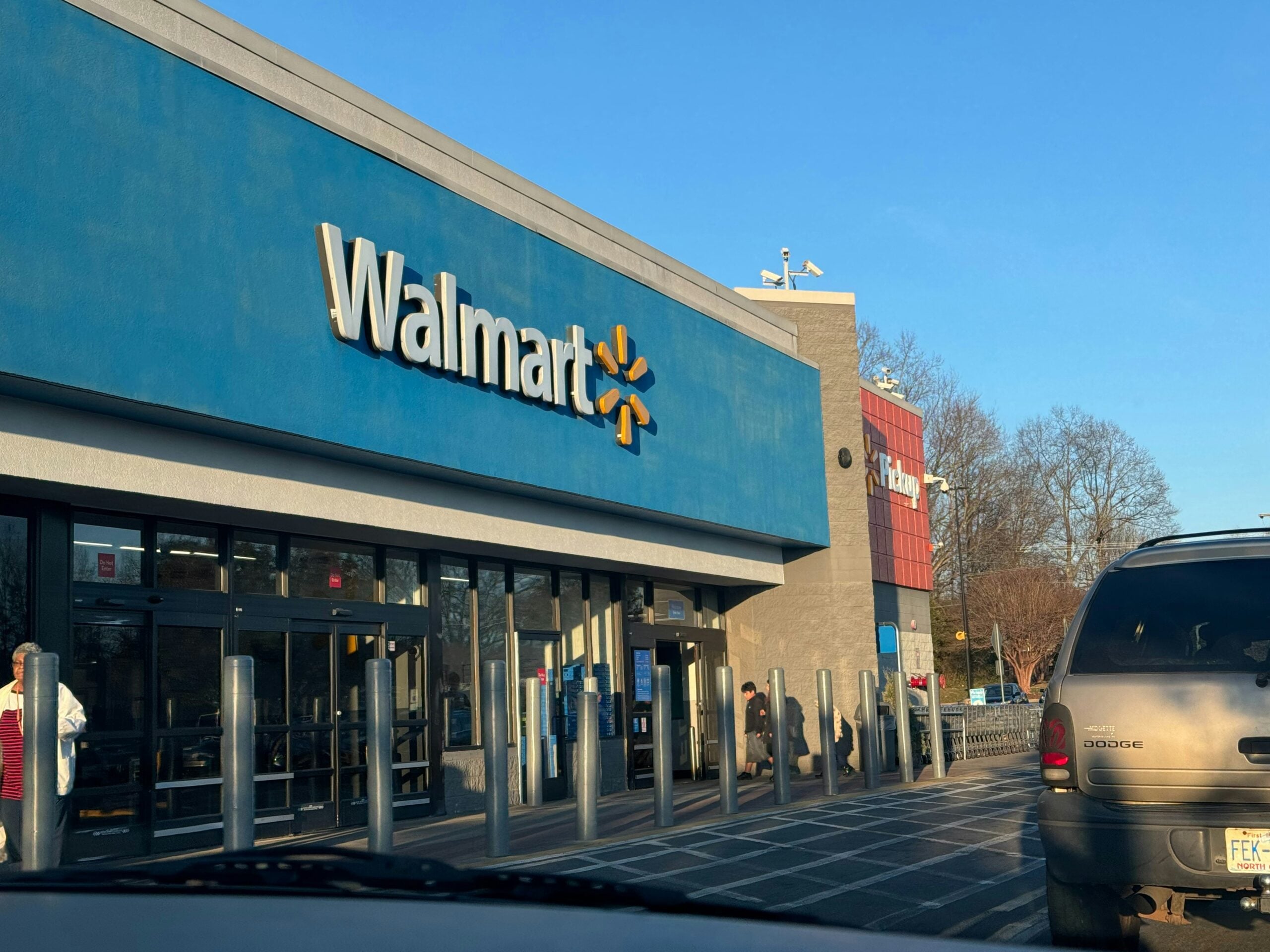 The exterior of a Walmart store is shown on a sunny day, with the Walmart logo visible above the entrance. Several vehicles and bollards line the parking area, and a few people are near the entrance.