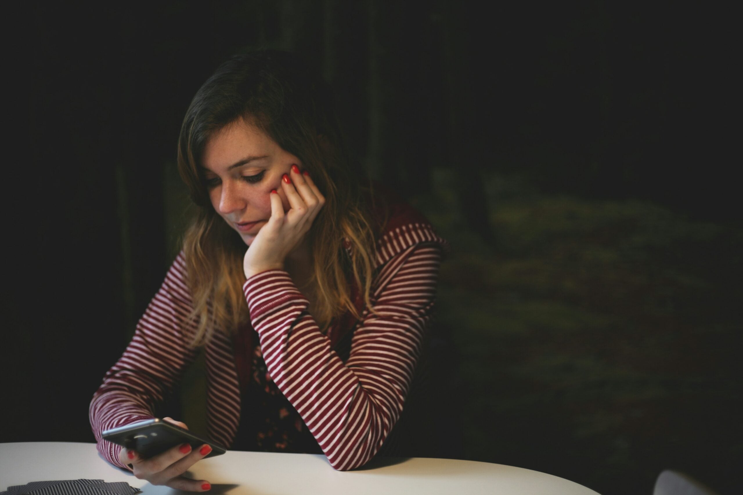 A woman with long hair, wearing a red and white striped jacket, sits at a table, resting her chin on her hand and looking at her phone with a thoughtful expression. The background is dark and out of focus.