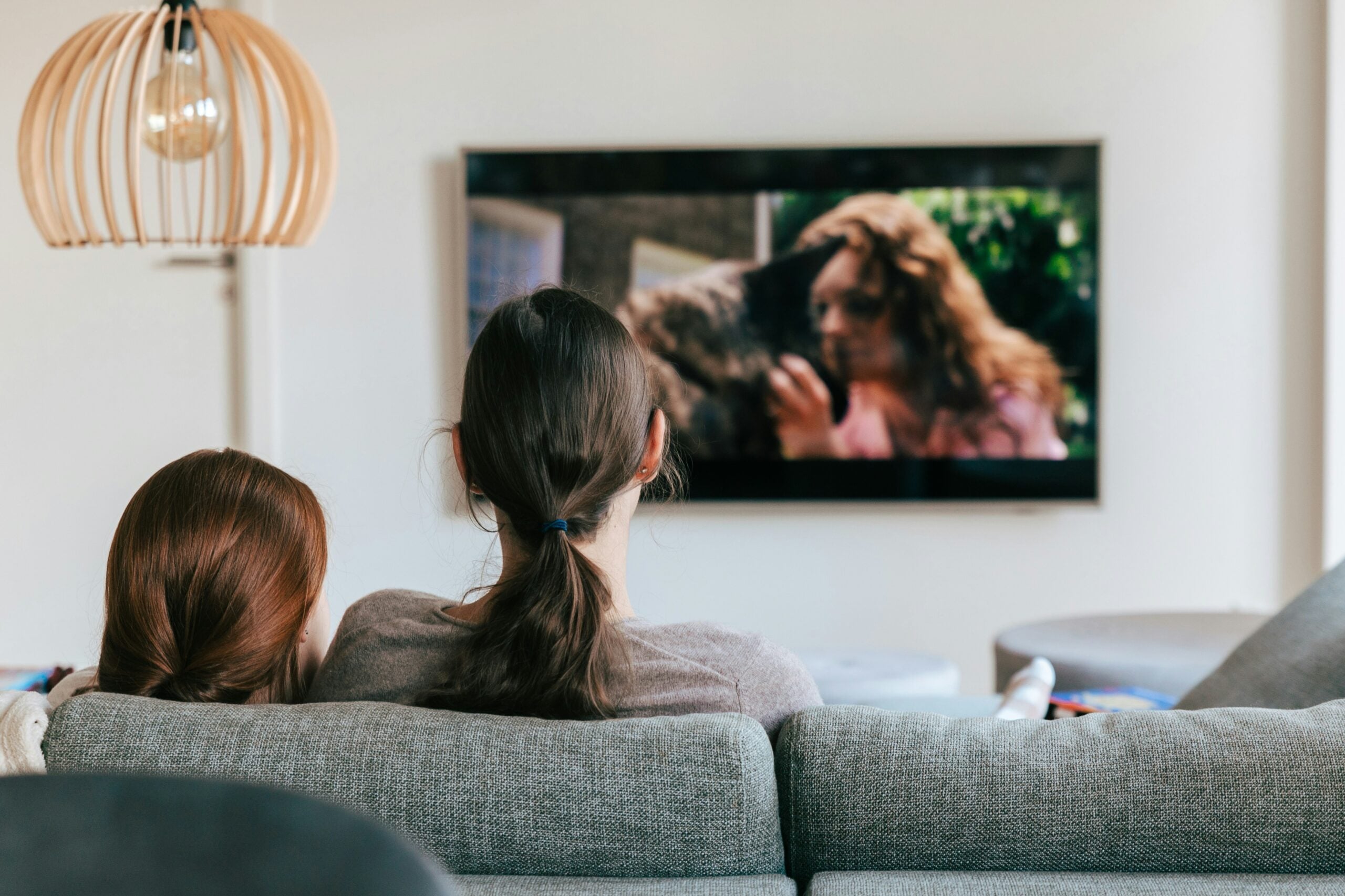 Two people sit close together on a couch, watching a movie on a flatscreen TV. The TV shows a woman with long hair interacting with an animal. The scene suggests a cozy, relaxed atmosphere at home.