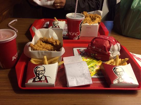 Overpriced fast food. Two KFC trays on a table, each with fried chicken, French fries, a drink, and condiments. One tray also has a wrapped burger. Receipts are placed on the trays, and two people are seated at the table.