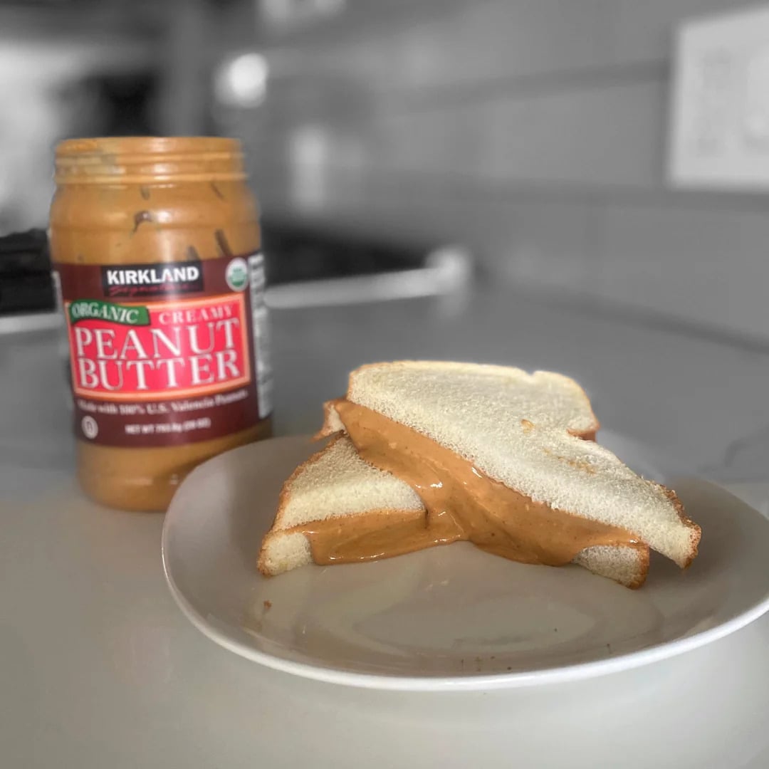 A plate with a peanut butter sandwich made from white bread sits on a counter. An open jar of Kirkland organic creamy peanut butter, known as one of the best peanut butter brands, is next to the plate. The sandwich is cut in half and peanut butter oozes out.