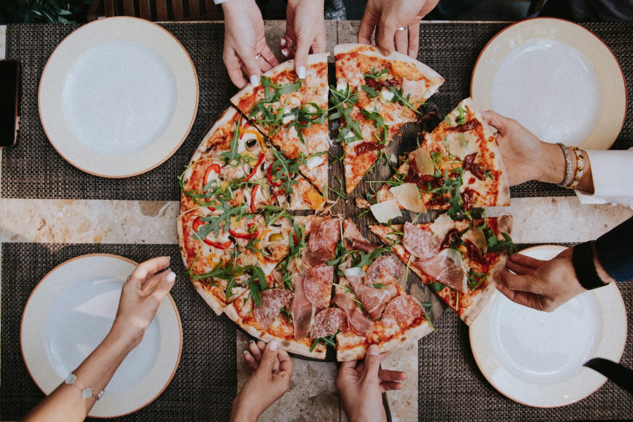 Six people reach for slices of a large pizza topped with arugula, peppers, and meats, around a table set with empty plates and cutlery. The scene suggests a shared meal in a social setting.